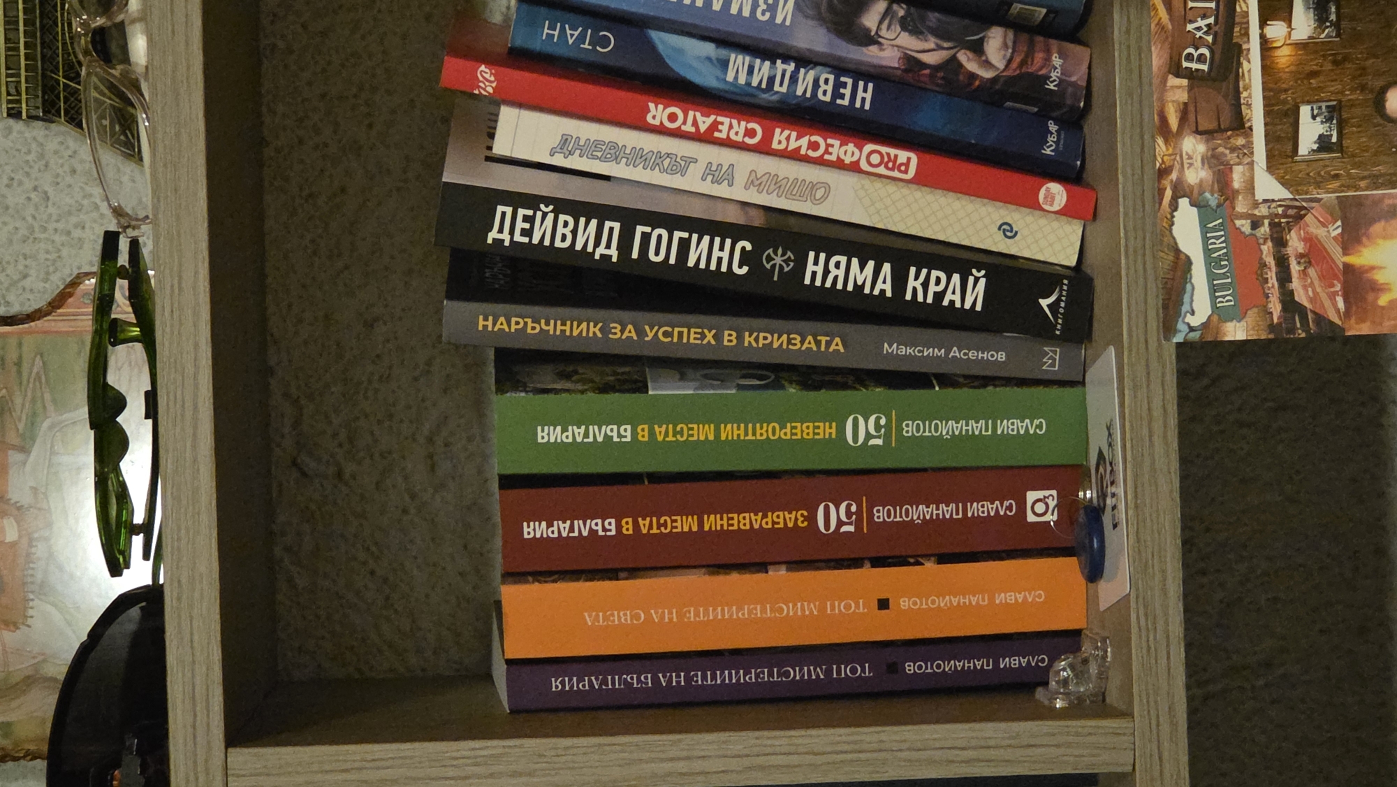 A shelf with several books and a pair of sunglasses is visible. The books are arranged vertically and have various titles and colored spines. The sunglasses are green and are placed on top of the shelf. Below the shelf, a postcard is visible, featuring an image of Bulgaria and a fireplace. The background appears to be a textured wall.