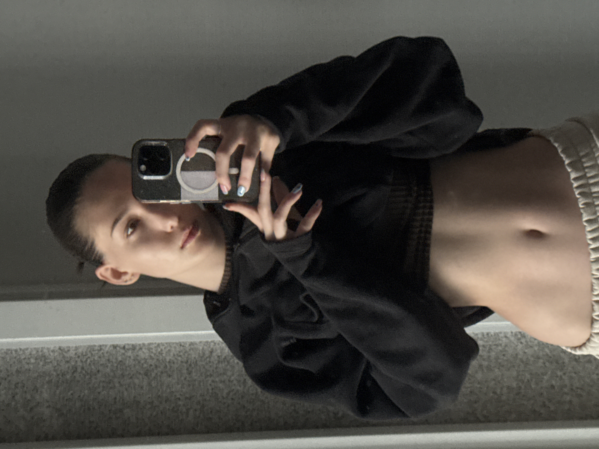 A young woman is taking a selfie in what appears to be an indoor location. She is wearing a dark, cropped sweatshirt and light-colored sweatpants. The background includes a wall and a carpeted surface. The woman is holding a smartphone with a decorative case. There is no information to ascertain if the location is Sofia, Bulgaria.