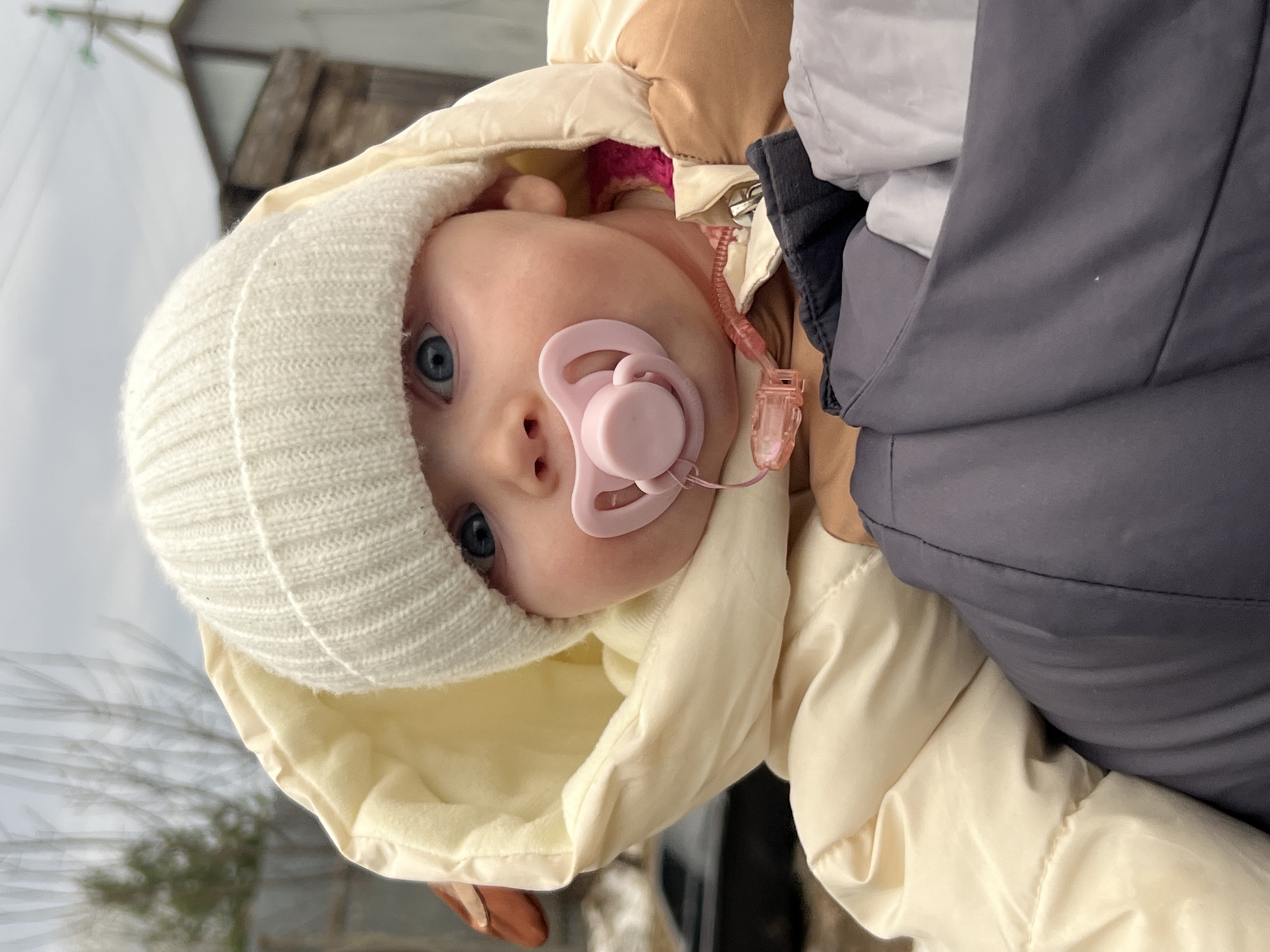 A baby with blue eyes and a pink pacifier is wearing a white knit hat and a cream-colored hooded jacket. The baby is facing the camera and is positioned against a background with a gray sky and a building structure in the upper left corner. The photo appears to be taken in an outdoor setting, possibly within the urban environment of Kamskoe, Russia, but this is based solely on the prompt's information and not evident in the image itself.