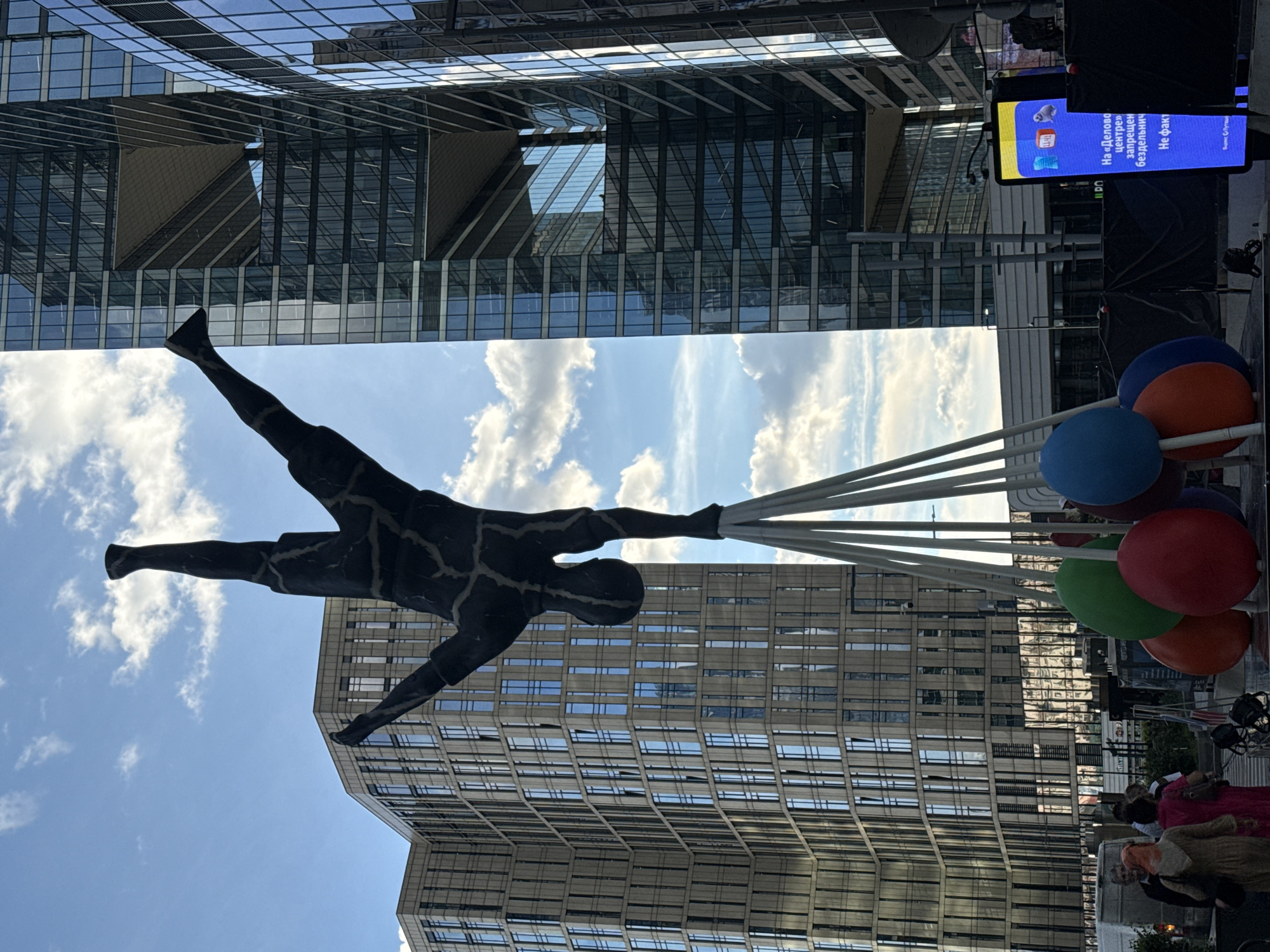 This low-angle, vertically oriented photograph captures a striking outdoor sculpture of a silhouetted human figure appearing to be lifted by a cluster of large, colorful balloons. The sculpture is dark, with a cracked or patterned texture on its surface. It is positioned in the foreground, with its arms outstretched, seemingly holding onto the strings of the balloons. The balloons are a vibrant mix of blue, red, green, and orange.
The scene is set against a backdrop of modern, glass-fronted buildings that reflect the sky and surrounding environment. The architecture suggests an urban setting with clean lines and geometric forms. The sky above is a bright blue, dotted with fluffy white clouds, indicating a pleasant day.
In the lower right portion of the image, a digital screen displays text in Russian. The visible text reads: "На «Деловой» в центре запрещен бесконтактный Не факт." This translates to: "On 'Business' in the center, contactless is prohibited. Not a fact." The screen also features a logo that appears to be an "i" inside a speech bubble.
Below the screen and to the right, several people are visible, some appearing to be standing or walking, dressed in casual attire. Their presence suggests this is a public space, possibly a plaza or promenade. The overall atmosphere conveyed is one of urban modernity combined with a playful artistic element, under a clear, bright sky. The time of day appears to be daytime, likely late morning or afternoon, given the brightness of the sky and the quality of light. The location is identified as Khimki, Russia.