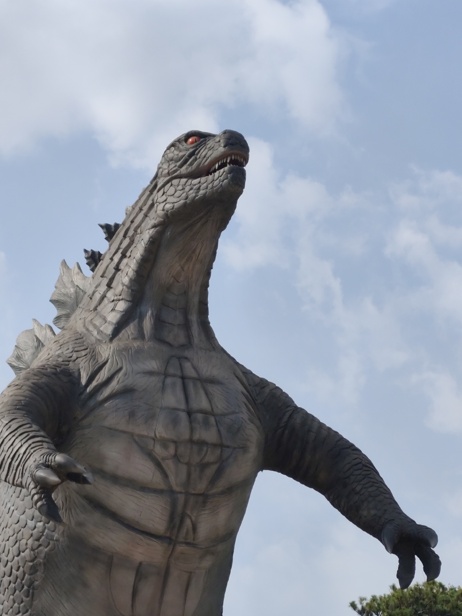 A low-angle shot captures a full-body view of a Godzilla statue. The statue is grey and textured, resembling reptilian scales. It stands with its head tilted upward, mouth slightly open, revealing teeth. The eyes are red. The statue's body is muscular, with prominent chest plates and dorsal fins along its back. The arms are extended outward, and one hand is visible with claws. The background is a light blue sky with scattered white clouds. At the bottom right, a small portion of a green bush is visible.
The statue is located in the city of Cam Ly - Đà Lạt, Vietnam.