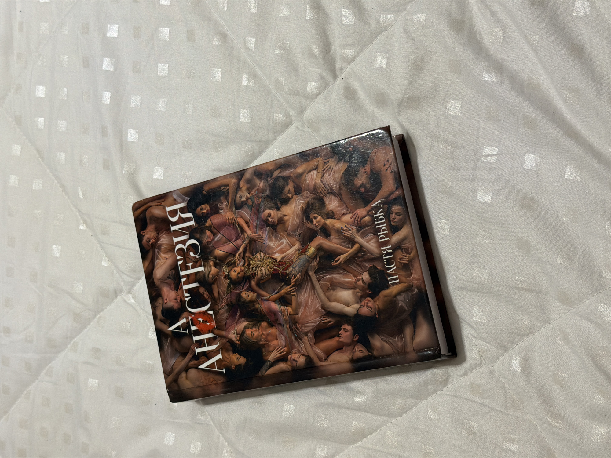 A book with a striking cover lies diagonally on a white, quilted surface. The surface features a subtle pattern of raised squares, suggesting a bedspread or blanket.
The book cover is rich in detail, depicting numerous intertwined human figures, both male and female, in a dynamic and intimate composition. Most figures appear to be unclothed or draped in light, flowing fabrics in shades of pink and gold. They are shown in various states of repose and embrace, creating a dense tableau of bodies. A central female figure stands out, adorned with elaborate gold and red embellishments, lying amidst the other figures. The overall style of the cover art resembles a classical painting with a dramatic, sensual, and perhaps dream-like aesthetic.
Visible text on the cover is in Cyrillic. Vertically aligned on the left side of the front cover is the title: "АНЕСТЕЗИЯ" (Anesthesia). The first "А" incorporates a distinctive red kiss mark symbol. On the right side of the cover, also aligned vertically, is the author's name: "НАСТЯ РЫБКА" (Nastya Rybka). The title "АНЕСТЕЗИЯ" is also partially visible along the spine of the book. The book appears to be glossy, reflecting some light.