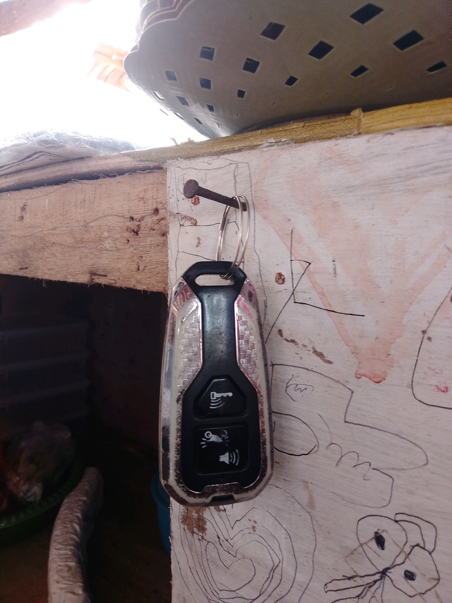 A silver and black key fob hangs from a nail on a rough, light-colored wooden surface. The key fob has three buttons, each with a symbol: a key, a car with sound waves emanating from it, and a car with headlights. The wooden surface appears to be part of a rustic structure, possibly a wall or partition. There are faint, childlike drawings on the wooden surface, including what appear to be stylized faces and abstract shapes, along with some red smudges. To the left of the key fob, a wooden beam is visible, and behind that, a dark, shadowed area contains what looks like a green bowl with some objects inside, one of which is long and light-colored and textured. The overall impression is an outdoor or semi-outdoor setting, possibly in a less-developed area, with some makeshift construction. The sunlight suggests it is daytime. The location is described as Cikal, Indonesia.