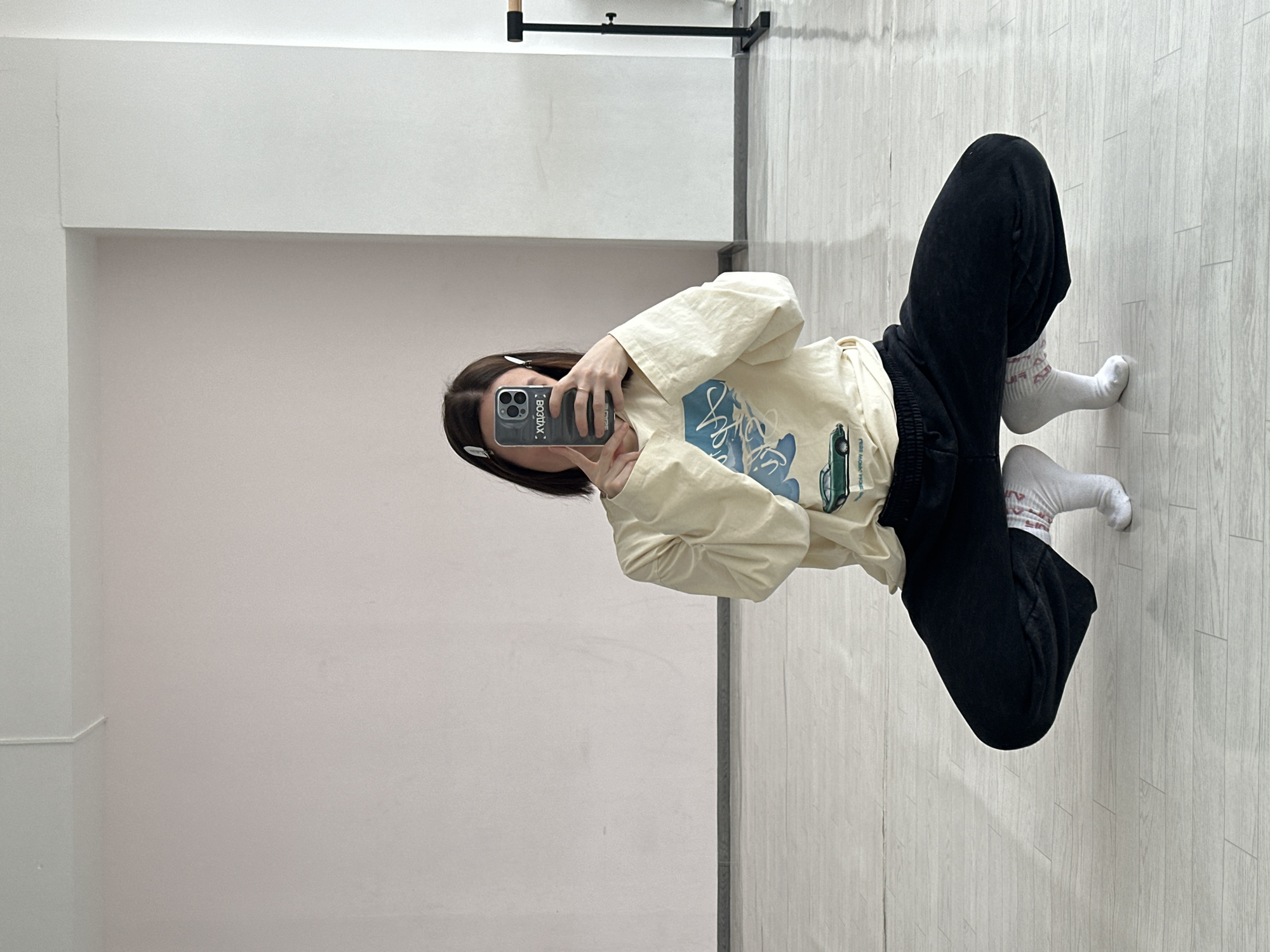 A young person is seated on a light grey floor in what appears to be a modern indoor space. They are wearing black sweatpants, white socks, and a cream-colored long-sleeved shirt with a graphic design on the front. The individual is holding a smartphone, taking a selfie. The phone case is dark and has writing on it. The person has dark hair with clips holding it back. The background consists of a plain white wall, with some sort of gymnastic training bar visible near the top of the frame. The overall impression is a casual selfie shot in an exercise or dance studio.
There is no evidence in the provided image to indicate that the location is Votkinsk, Russia.