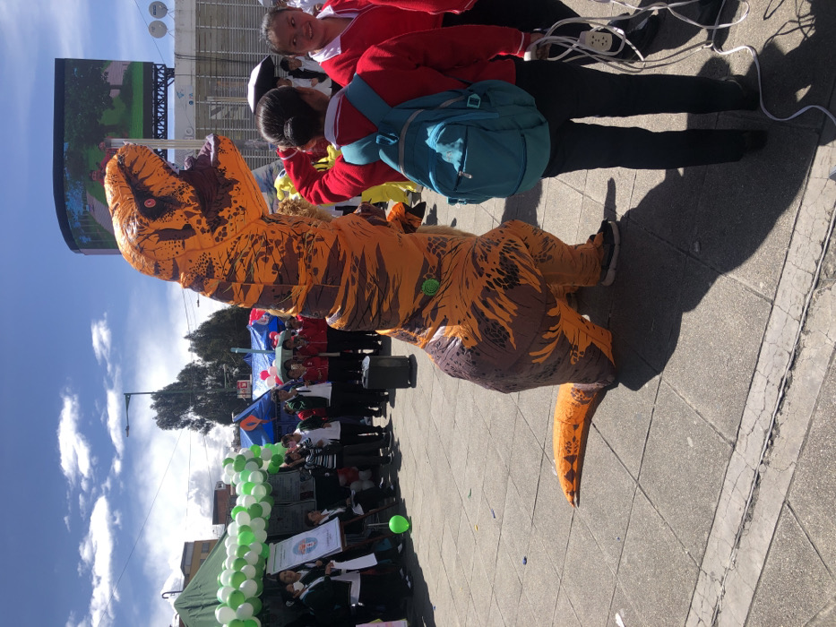 The scene depicts an outdoor public event taking place on a paved square in La Paz, Bolivia, under a partly cloudy sky. In the foreground and center, an individual in a full-body inflatable orange and brown Tyrannosaurus Rex costume stands upright, facing right with its mouth open. A small green circular object is affixed to the costume's lower back. To the immediate right, two young females are visible. One, facing away, wears a red long-sleeved top, dark pants, and a teal backpack. The other, partially visible, also wears a red top and holds coiled white electrical cords with a power strip in her left hand.
To the left of the T-Rex, a group of people is gathered under a temporary canopy decorated with green and white balloons. One person is speaking into a microphone while facing the group. A black portable speaker on wheels rests on the ground near the speaker. An easel displaying a white sign stands within this gathering. A single green balloon lies on the paved ground near the canopy. In the background, additional individuals are visible around other temporary stalls covered by blue and red canopies. A large digital billboard displaying an image of green trees is mounted on a building in the far background. The ground is a light-colored paved surface with visible cracks.