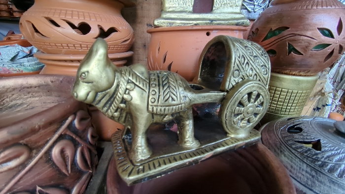 The image displays a close-up view of handicrafts, situated within what appears to be a market stall or shop in Dhaka, Bangladesh. The primary subject is a golden-colored metallic figurine of an ox pulling a two-wheeled cart, positioned centrally. The ox features embossed decorative patterns across its body, including geometric shapes and simulated harness straps. The cart possesses ornate side panels and spoked wheels. The figurine rests on a rectangular base with a patterned border.
In the background and around the central figurine, various terracotta and earthenware pottery items are visible. These include several unglazed brown pots and vases, some featuring cut-out designs such as heart shapes and leaf motifs, and others exhibiting embossed patterns. One prominent pot has a metallic gold-painted base with a distinctive grid texture, while another partially visible item on the right side features a silver-painted lid with intricate cut-outs and designs. No people or active interactions are depicted; the scene is a static arrangement of merchandise.