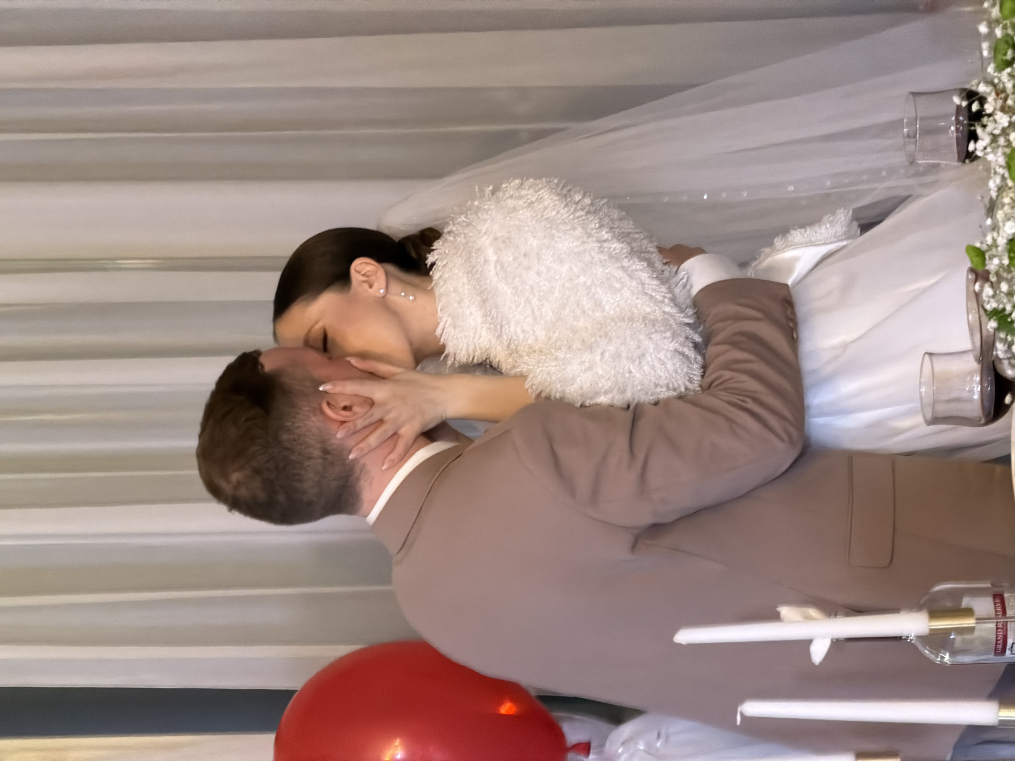 A wedding photograph shows a bride and groom kissing. The bride, wearing a white wedding dress, a white veil, and a fluffy white shawl, is embracing the groom. The groom is wearing a light brown suit. The scene includes wine glasses, candles in wine bottles, and a red balloon. The photograph was taken in Chaikovsky, Russia.