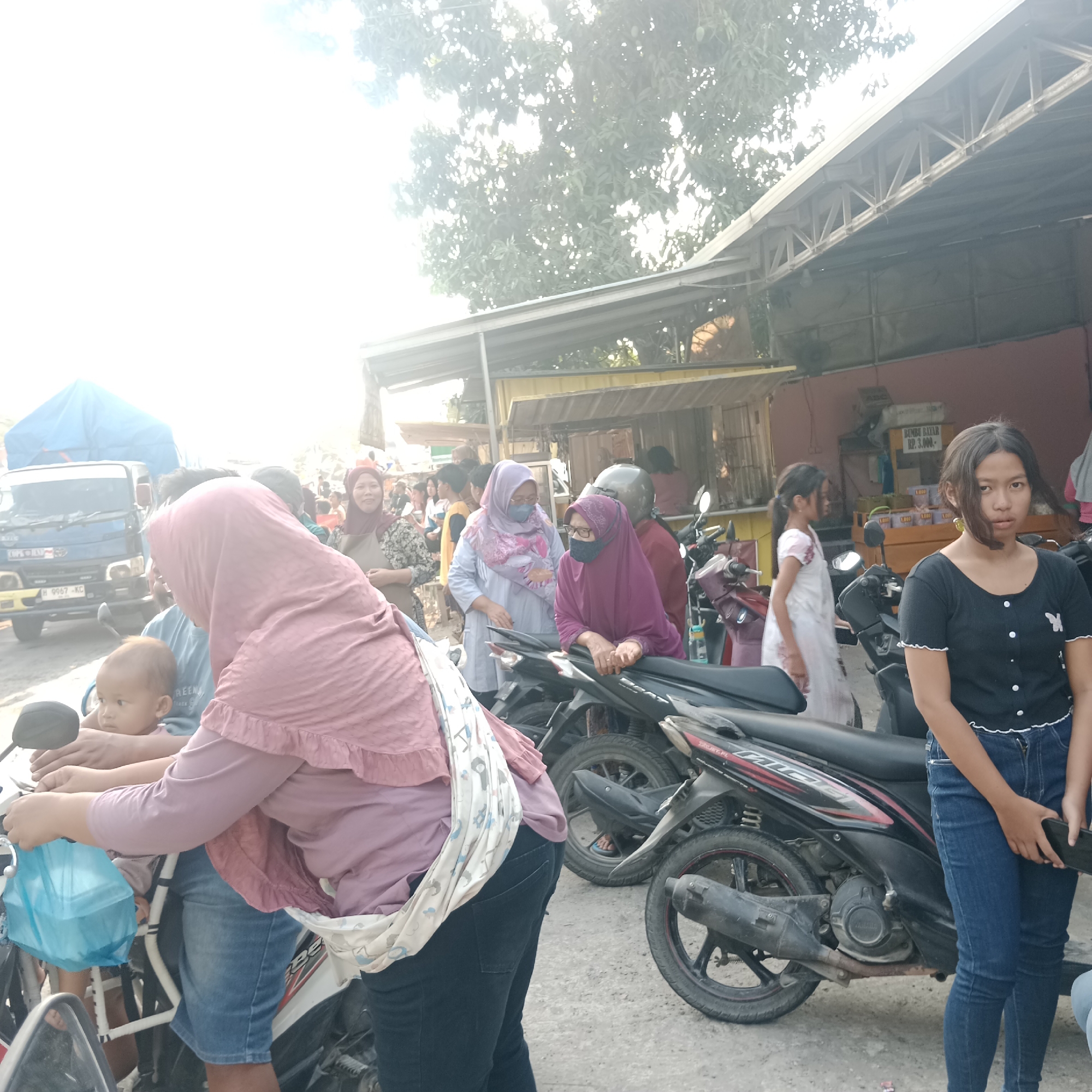 The image depicts an outdoor scene, likely a street or market area, in Tegalrejo, Indonesia, under bright, possibly hazy sunlight. A blue truck with a license plate H 9967 KC is parked on the left. In the foreground and midground, several people are visible, many of whom are wearing headscarves.
A woman in a pink headscarf and a white and patterned sling carrier, holding a baby, is seated on a motorcycle. The baby, facing left, has short dark hair. To her right, a woman in a maroon headscarf and glasses is leaning on a motorcycle. Another woman in a light purple headscarf and glasses is standing nearby. Further back, a young girl in a white dress with floral accents is walking. On the far right, a teenage girl with long dark hair, wearing a black short-sleeved shirt and blue jeans, is looking directly at the camera, holding a dark object in her hands.
Several motorcycles are parked in the scene, some with passengers. In the background, there are stalls with awnings and signage, including one that reads "BOMBO BAYAR RP. 3.000,-". The environment appears to be a busy public area, possibly a roadside market. The overall lighting suggests it is daytime.