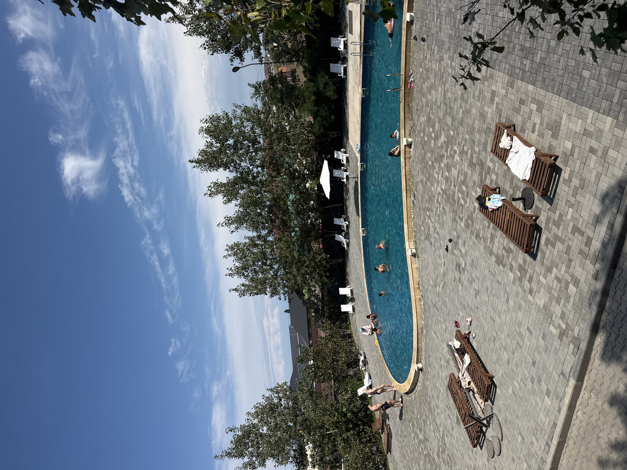 The image captures an outdoor swimming pool area in the city of Akshi, Kazakhstan, on a clear, sunny day. The pool, filled with bright blue water, has a kidney shape with a curved edge. Several people are visible in and around the pool. In the water, individuals are swimming and standing. Along the poolside, there are lounge chairs made of dark wood, some with towels or bags on them. A white umbrella provides shade for a cluster of lounge chairs in the background. The ground surrounding the pool is paved with interlocking gray bricks. Lush green trees with full canopies are situated behind the pool area, with buildings and a fence visible in the distance. The sky is a vibrant blue with scattered white clouds.