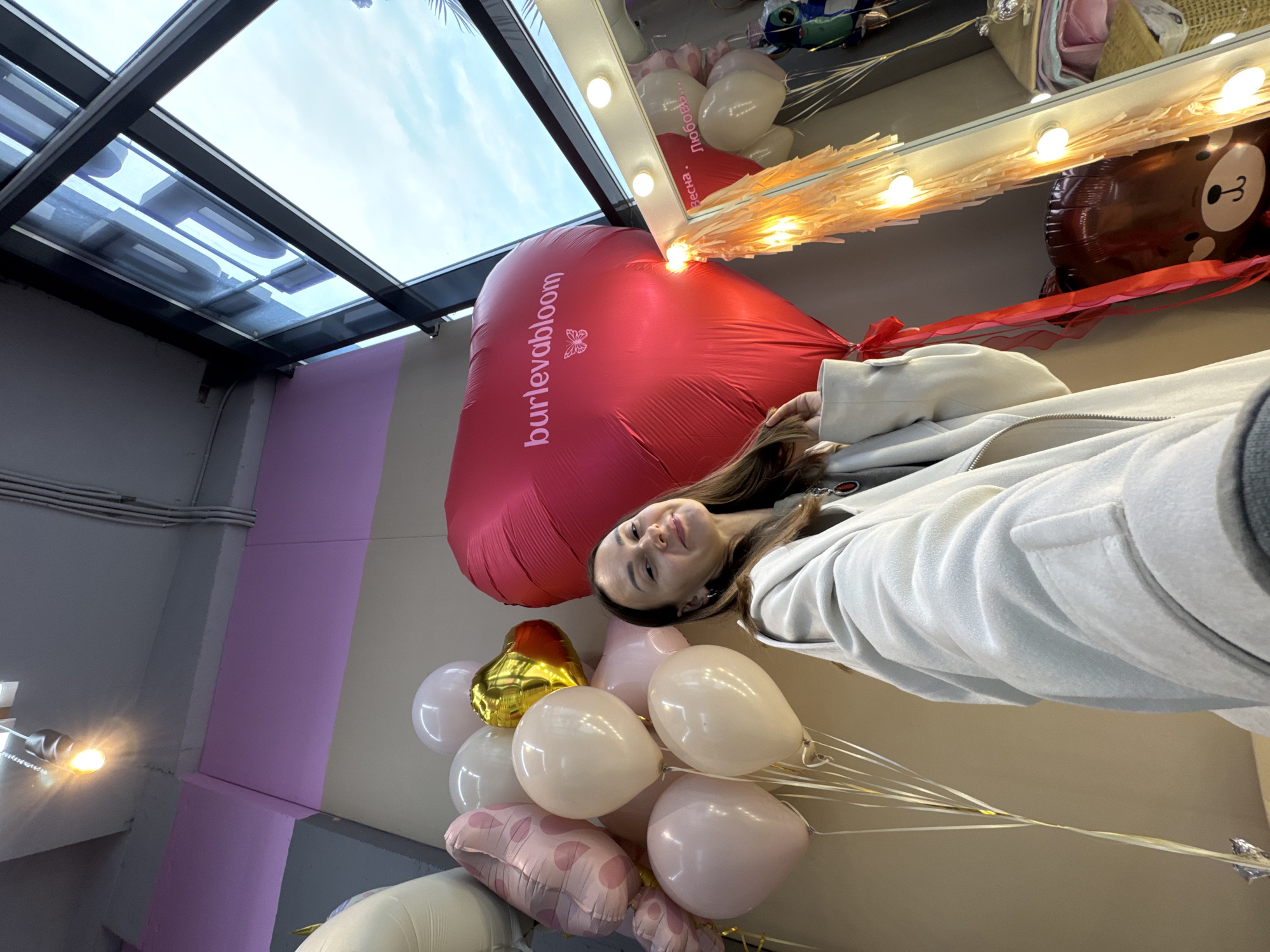 A young woman is posing indoors with a large red heart-shaped balloon with the text "burlevabloom" printed on it and several other balloons of different colors (white, gold, pink) in what appears to be a store. She is wearing a light-colored coat. The background includes a mirror with lights and other balloons and decorations. The lighting in the store is bright. The ceiling has a modern skylight. The image is likely taken in Sochi, Russia, based on the text on the balloon.