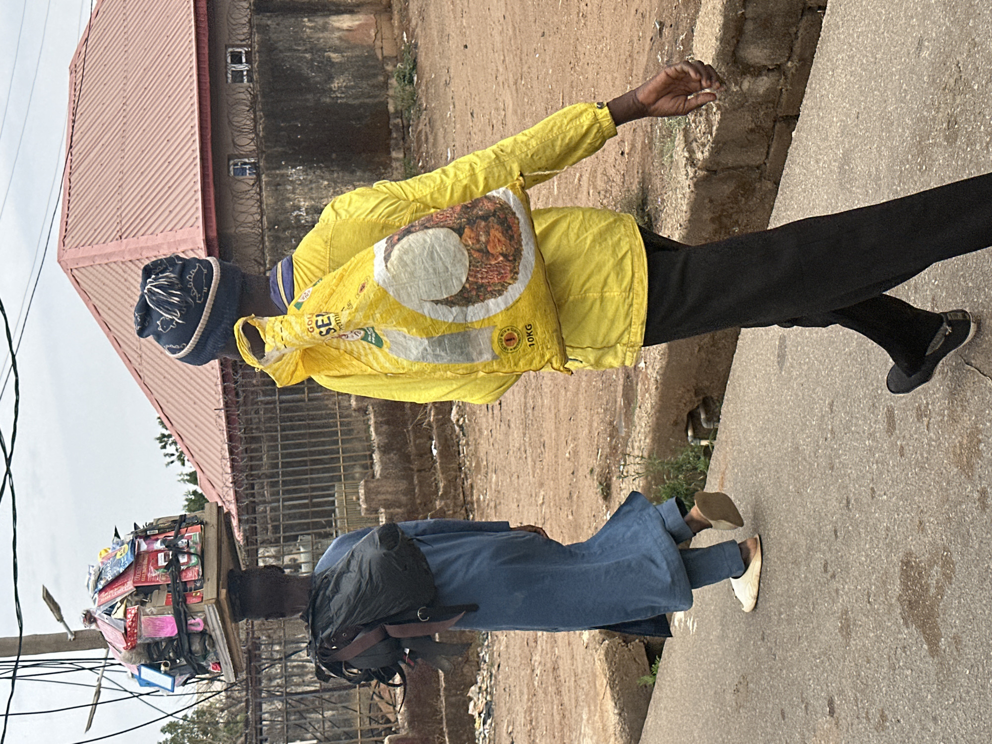 The image, rotated 90 degrees clockwise, captures a street scene in Jos, Nigeria, featuring two individuals engaged in everyday activities. The setting appears to be a paved sidewalk alongside a dusty or unpaved area, with a building in the background under an overcast sky.
In the foreground (left side when rotated), a person walks, dressed in a bright yellow long-sleeved jacket, black trousers, and dark shoes. They wear a dark knitted cap with white patterns. Over their left shoulder, they carry a large yellow bag. The bag prominently features an image of food, likely rice and stew, and bears the visible text "GOLDEN SEA" and "10KG", suggesting it is a bag of rice. Their right arm is slightly extended.
Further back (right side when rotated), another person walks, wearing a long, dark blue garment, possibly a traditional thobe, and light-colored slip-on shoes. They have a black backpack on and are balancing a large, rectangular wooden tray or box on their head. This tray is heavily laden with various small packaged goods, boxes, and items that appear to be stationery or small household products, indicative of a street hawker.
The background includes a building with a reddish-brown corrugated metal roof and concrete walls. Barbed wire is visible along the top of a wall, and small windows are visible on the building's concrete facade. Overhead utility lines are also visible on the far left of the frame. The overall impression is a bustling street during daytime, with people going about their daily routines, including commerce.