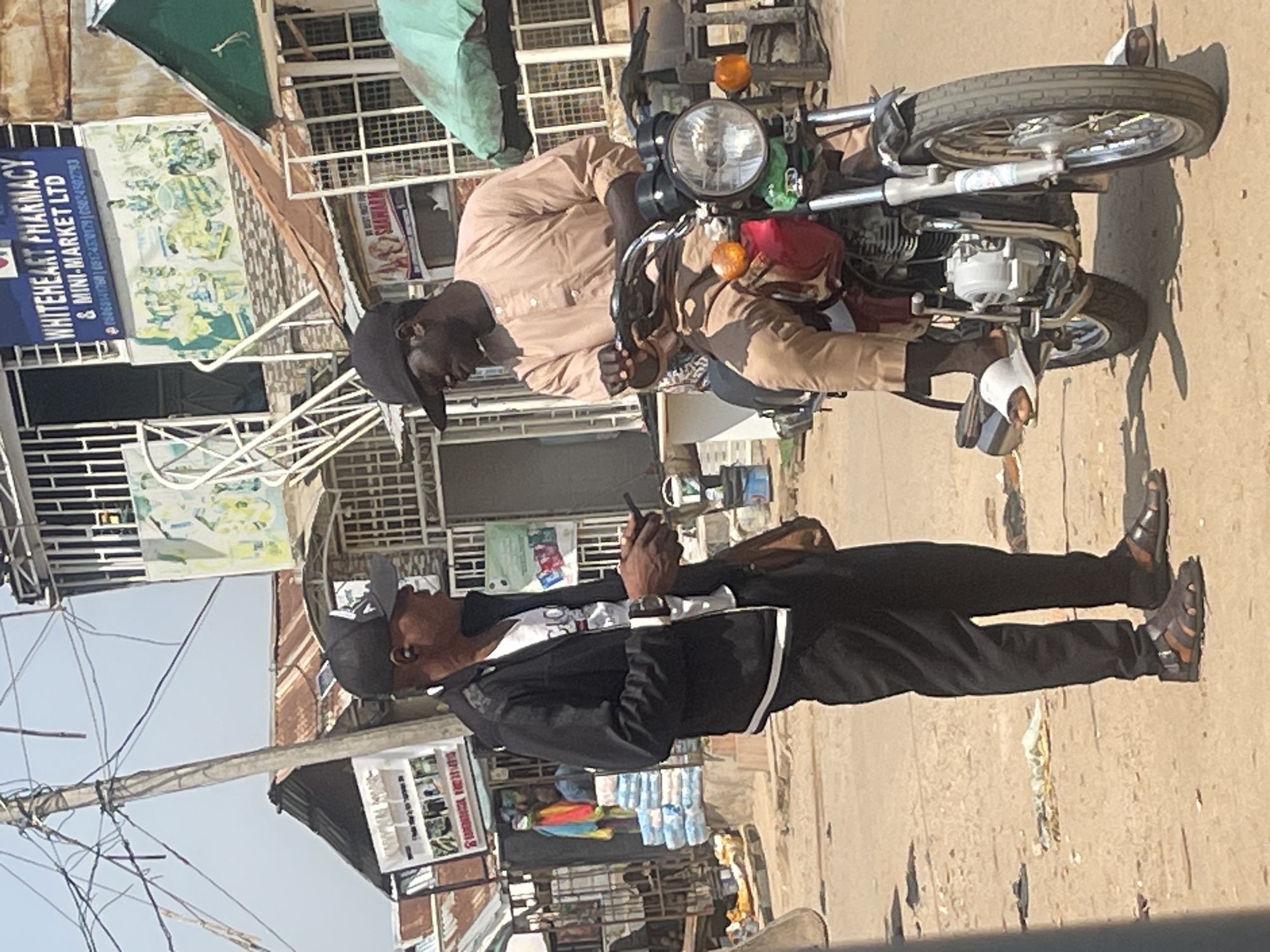 The image depicts a street scene in Abuja, Nigeria. Two men are the primary subjects. The first man is sitting on a motorcycle, wearing a light brown traditional outfit and a black cap. He appears to be holding something in his right hand. The second man is standing beside the motorcycle, facing away from the viewer. He is wearing a black jacket over a white t-shirt and black pants, also with a black cap. He is looking down at a mobile phone he is holding in his right hand.
The motorcycle appears to be a standard commuter bike with a prominent headlight and turn signals. In the background, there are buildings with storefronts. One storefront has a sign that reads "WHITEHEART PHARMACY & MINI-MARKET LTD". There are also other signs and posters visible, though much of the text is difficult to decipher. Shelves stocked with various items, including what appear to be bottled water and colorful clothing, are visible in the background. Overhead, there are numerous electrical wires crisscrossing the sky, and some awnings or tarps are extended from the buildings.
The lighting suggests it is daytime, with shadows cast on the ground, indicating the sun is likely out. The overall scene conveys a busy, everyday atmosphere of a commercial area in a city.