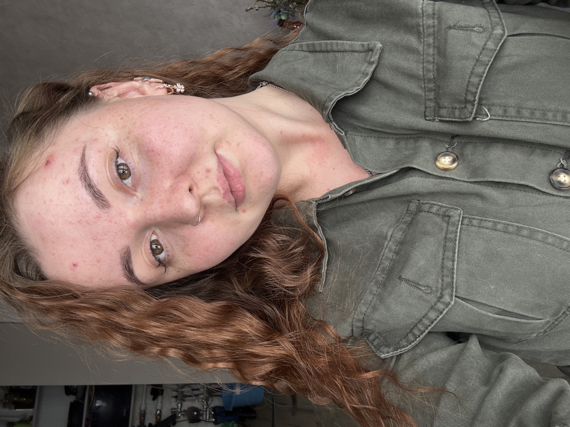 A woman with long, auburn, curly hair is wearing a dark green, collared, button-down shirt. She has fair skin with freckles. She has an earring in her left ear, a nose ring in her left nostril, and light brown eyes. The background is a light gray color. The photo is taken at a slight angle. There is nothing to suggest the location is Фоки, Russia.