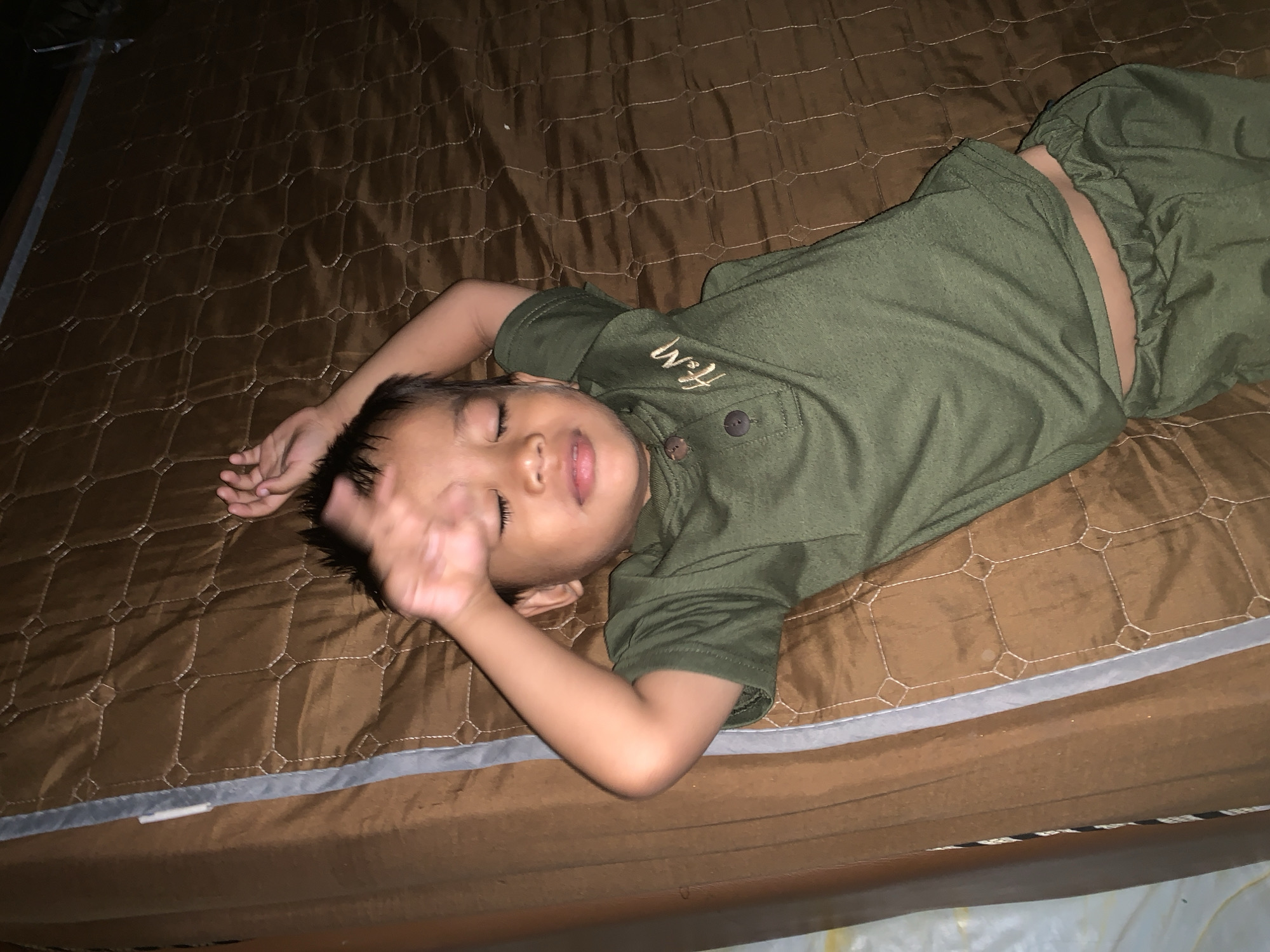 A young child lies on a brown, quilted bed with a silver-gray border. The child is wearing green clothing, including a short-sleeved shirt with the letters "H&M" embroidered on the chest and matching pants. The child's eyes are closed, and their right hand is raised near their face. The image was taken indoors, likely in a residential setting in Sungai Meriam, Indonesia.