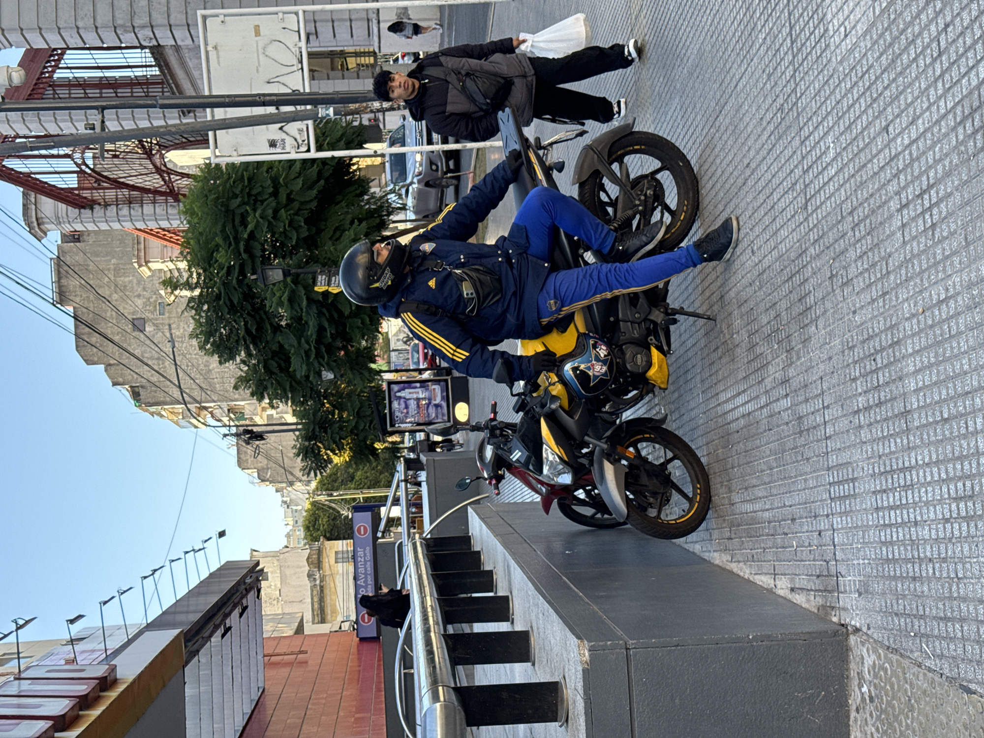 A medium shot captures two men with motorcycles in a Buenos Aires, Argentina street scene. One man, dressed in a blue and yellow tracksuit and a helmet, is seated on a yellow and black motorcycle. The second man, in a dark jacket and pants, stands nearby, holding a white plastic bag. Two motorcycles are parked on the sidewalk, which is made of square gray tiles. In the background, a building facade with a red metal structure is visible, along with trees, street lights, and other buildings. A stairway leading to a lower level is located next to the motorcycles.