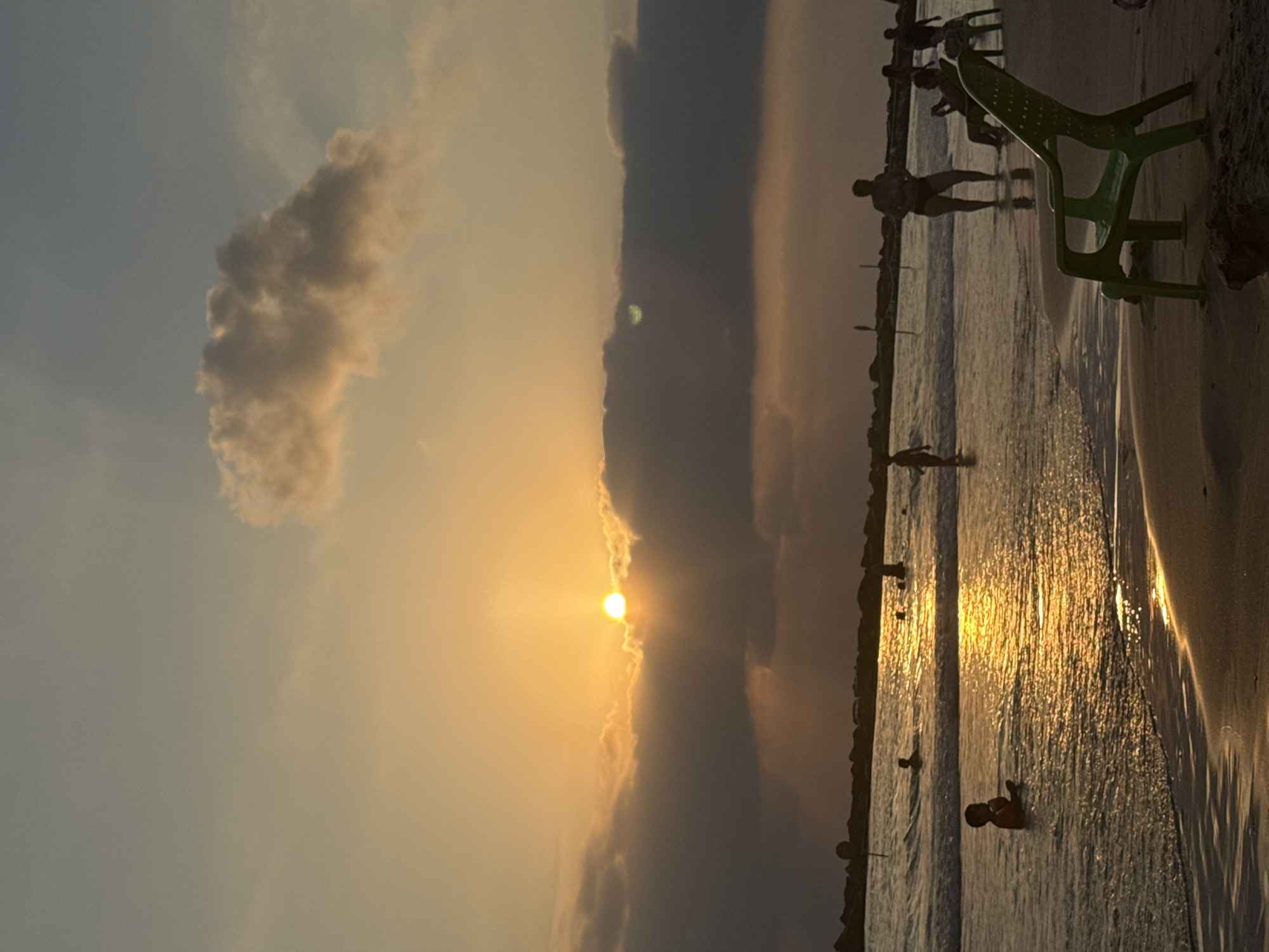 The image captures a sunset at a beach in Netanya, Israel. The sun is low on the horizon, partially obscured by dark clouds, casting a warm golden light across the sky and water. The sea is calm with gentle waves lapping at the shore.
Several people are visible on the beach and in the water. In the foreground, a green plastic beach chair sits on the sand. Further back, a child is playing in the shallow water. Other individuals, silhouetted against the bright sky, are walking along the beach or wading in the sea. Some are in swimwear. A line of dark rocks or a breakwater is visible along the edge of the water in the distance, with some structures, possibly lifeguard posts or poles, extending upwards. The overall atmosphere is serene and peaceful, typical of a beach at sunset.