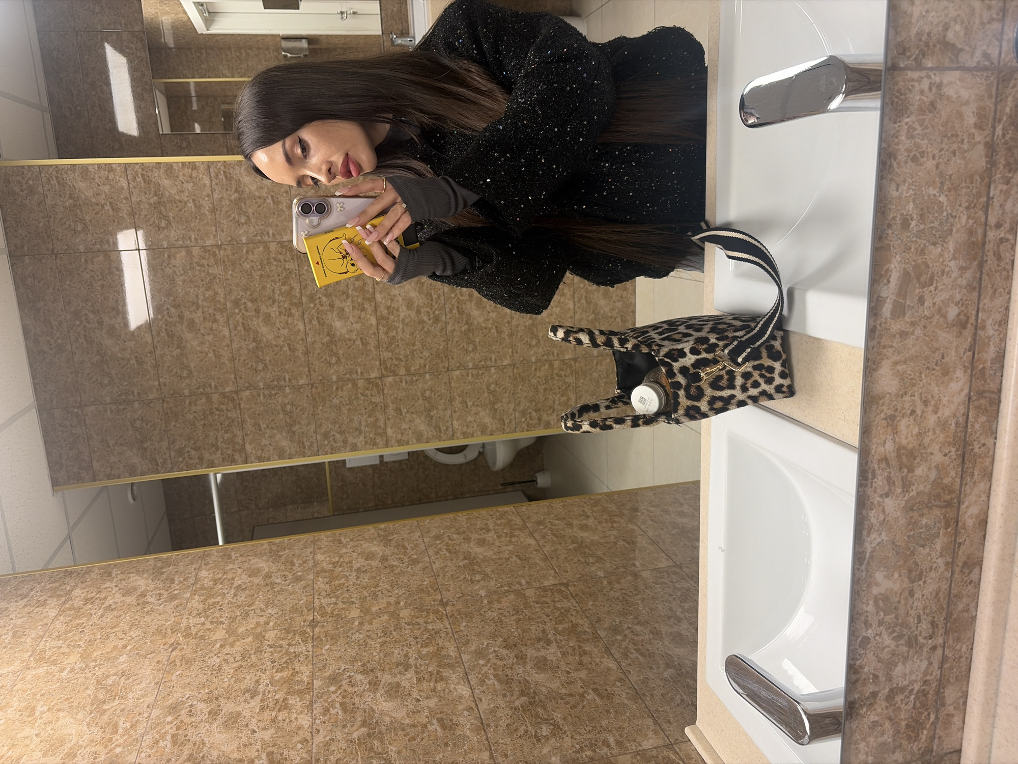 A person is taking a selfie in a bathroom with brown tiled walls, holding a cell phone with a yellow case. The person is wearing a black jacket and grey fingerless gloves. A leopard print handbag is on the counter near the sink. The location is a bathroom with a white sink and a brown-tiled wall in Saint Petersburg, Russia.