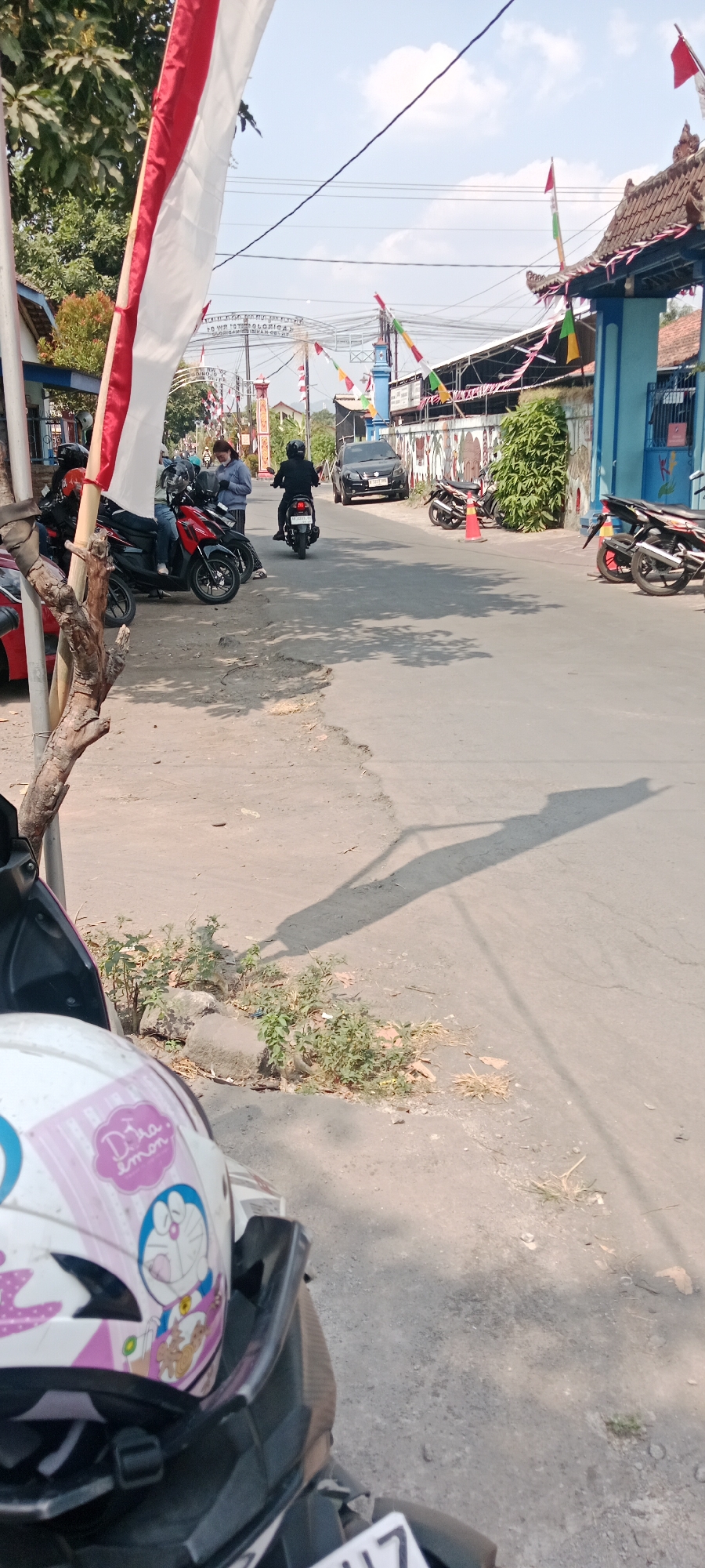 The image captures a street scene in Purwomartani, Indonesia. In the foreground, the front of a motorcycle with a white helmet decorated with Doraemon and pink accents is visible. The helmet is angled slightly to the left. To the left of the motorcycle, a wooden pole with a flag partly obscuring it stands. A large shadow of a pole is cast on the road.
In the middle ground, several motorcycles are parked along the left side of the road. Two people are interacting near the parked motorcycles; one is wearing a blue jacket and is looking at their phone, while the other, wearing a black jacket and helmet, is on a motorcycle. Further down the road, a person in a black jacket and helmet is riding a motorcycle away from the camera. A black car is also visible in the middle ground.
The environment is characterized by a paved road with uneven edges and patches of dry grass and small plants growing along the roadside. The background features a building with a tiled roof and blue entrance gates, decorated with Indonesian flags and colorful pennants. Power lines are visible crisscrossing the sky. The overall lighting suggests a sunny day with a clear sky and scattered clouds.