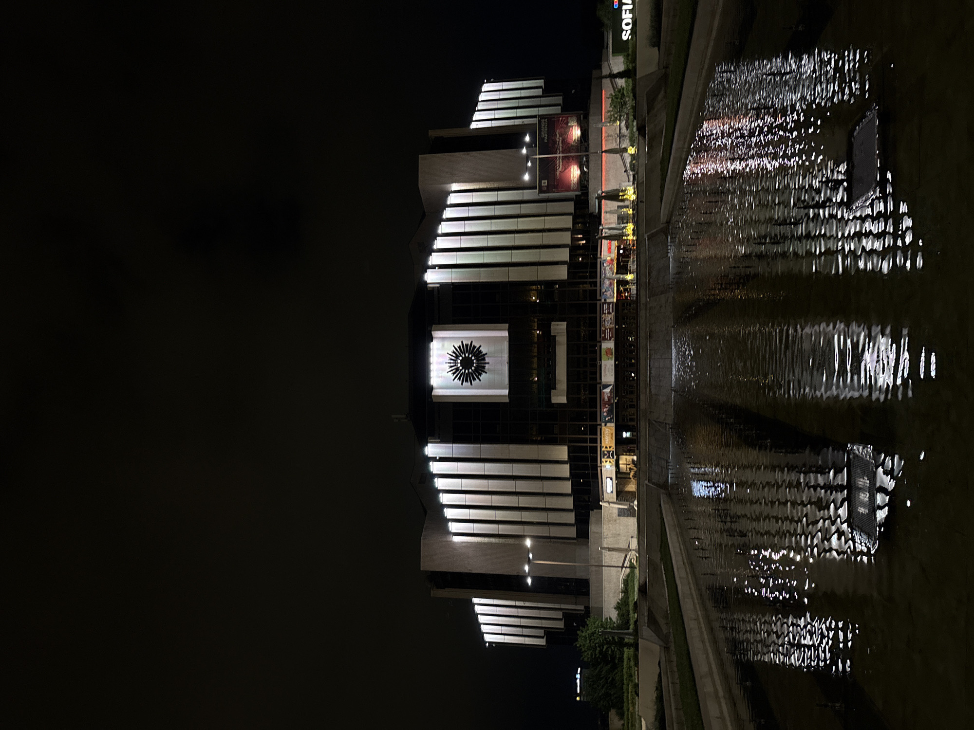 The image depicts the National Palace of Culture (NDK) in Sofia, Bulgaria at night. The building is a large, modern structure with illuminated horizontal white panels on its facade, reflecting in the dark water feature in the foreground. The reflection shows the structure and its lights. The night sky is dark, and the surrounding area is dimly lit, with signs indicating "SOFIA". Vegetation is visible on the right side of the image. No people are visible.