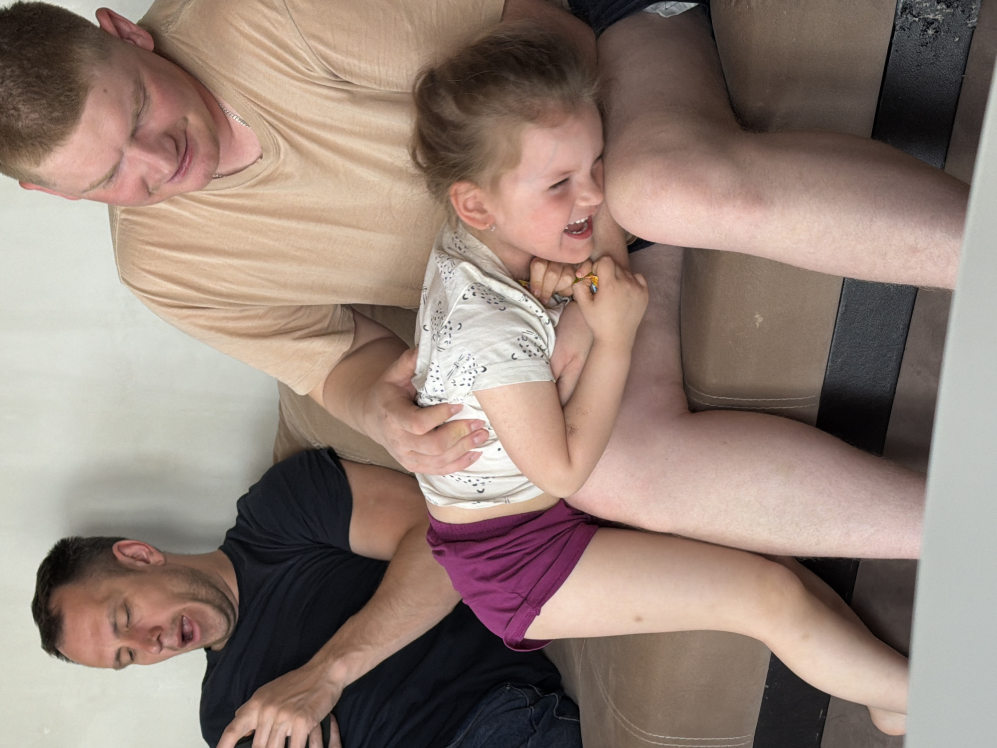 The image depicts a Caucasian child with light brown hair, wearing a white short-sleeve shirt with dark patterns and maroon shorts, sitting on the laps of two adult males. The child is smiling and appears to be laughing. One of the men is wearing a tan t-shirt and appears to be looking at the child with affection. The other man is wearing a black t-shirt and has his mouth open, possibly laughing or talking. They are situated on a brown and black couch. The setting might be in an apartment or home based on the couch and plain wall. The location provided is Chaikovsky, Russia, however, there is nothing visible in the image to confirm that location.