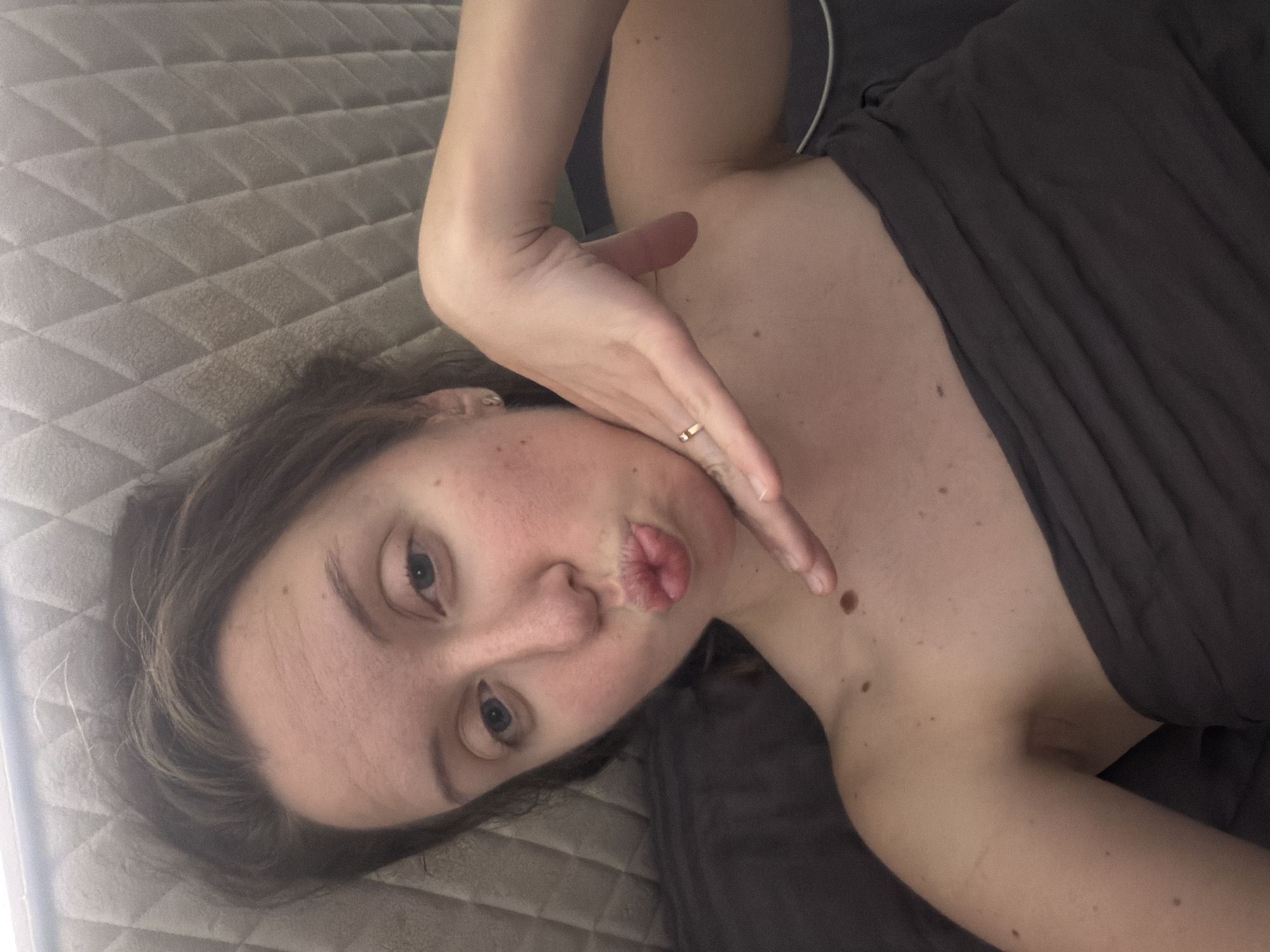 A woman with brown hair and blue eyes is lying on a quilted surface, likely a bed or couch, and looking into the camera. She is wearing a dark gray strapless top. Her right hand is raised to her face, with her index finger resting near her chin. She is puckering her lips. There are several moles visible on her left shoulder and upper chest. The background is a gray quilted fabric, and a dark gray surface is visible beneath her. There is no indication of the city of Bila Tserkva, Ukraine, in this image.