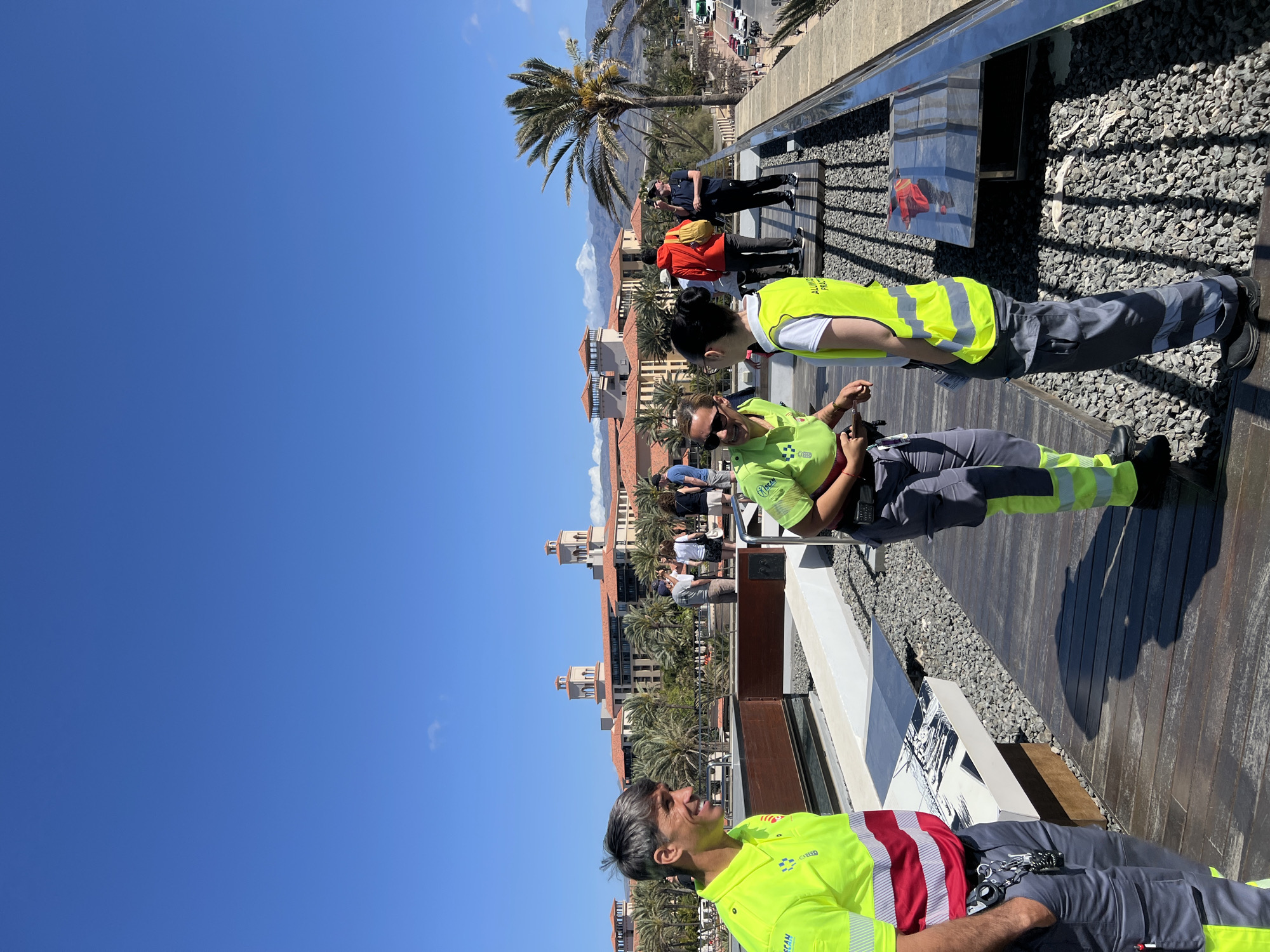 The image shows three individuals wearing neon yellow vests with gray pants, standing on a wooden deck or platform in what appears to be an outdoor setting. The city of Maspalomas, Spain, is visible in the background, including buildings with red tile roofs and palm trees. The sky is clear and blue. The individuals appear to be engaged in conversation. Other people can be seen in the background. The scene is well-lit, suggesting a sunny day. Part of the platform has gravel and a metal railing.