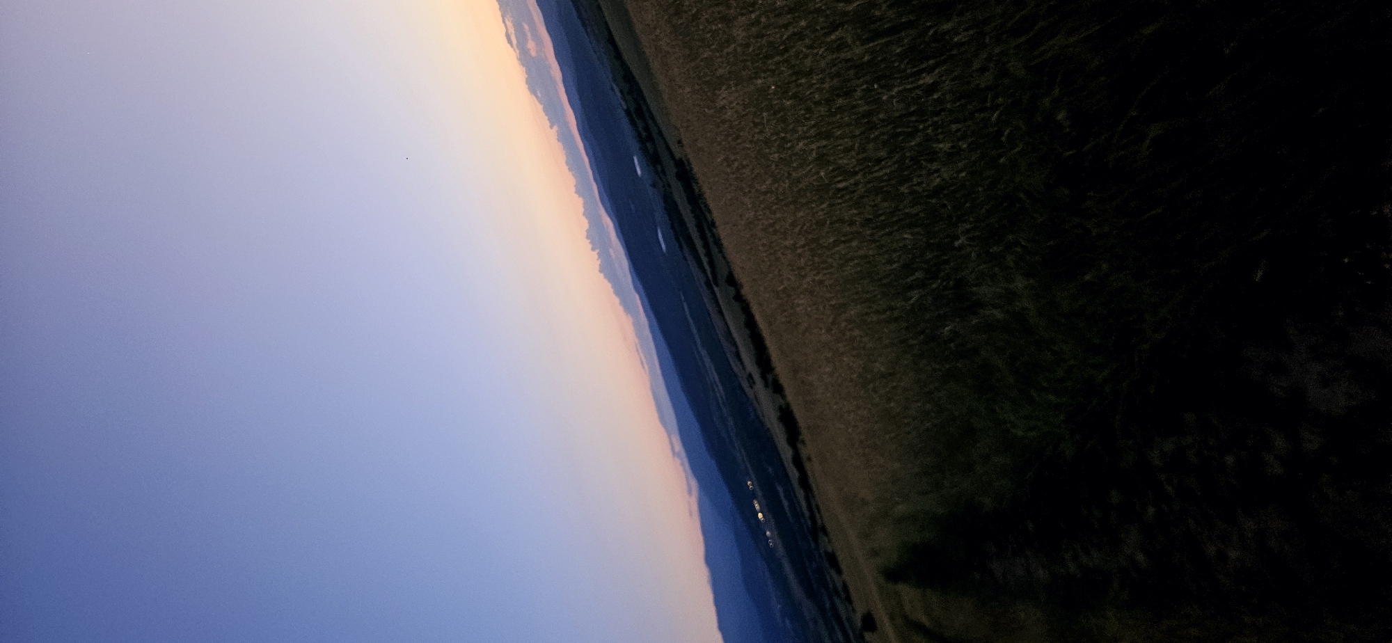 The image shows a landscape view of the area near Momin Prohod, Bulgaria, during twilight. A vast field of tall grass dominates the foreground, appearing dark and somewhat blurred. A dirt path or track is visible on the left side of the field, also mostly in shadow. In the background, rolling hills and mountains are silhouetted against a twilight sky. The sky transitions from a deep blue at the top to lighter shades of purple and orange near the horizon, indicating the presence of sunset or sunrise. Some faint lights, possibly from a settlement, can be seen in the distance on the slopes of the hills. The overall impression is of a rural, natural setting at the edge of day.