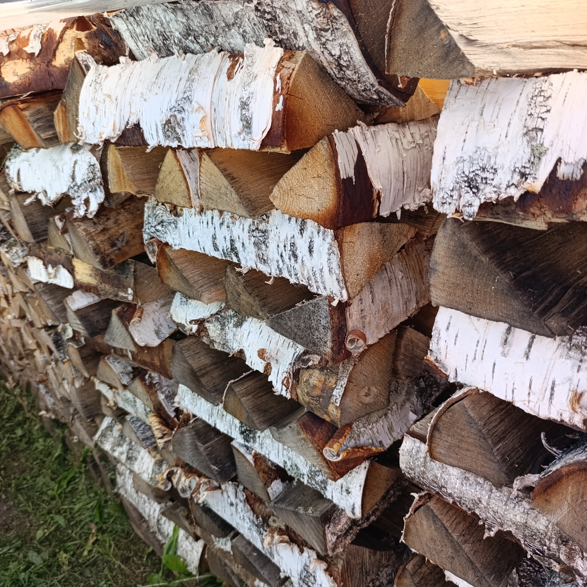 The image depicts a neatly stacked pile of chopped firewood, primarily composed of birch logs, with their characteristic white and dark bark. The logs are cut into various lengths and arranged in horizontal layers. The firewood is stacked outdoors, with a grassy area visible in the lower left portion of the frame. The scene appears to be a yard or storage area in the city of Dubovaya, Russia, likely prepared for winter use. There are no people or animals present in the image. The lighting suggests it is daytime, possibly late afternoon given the warm tones.