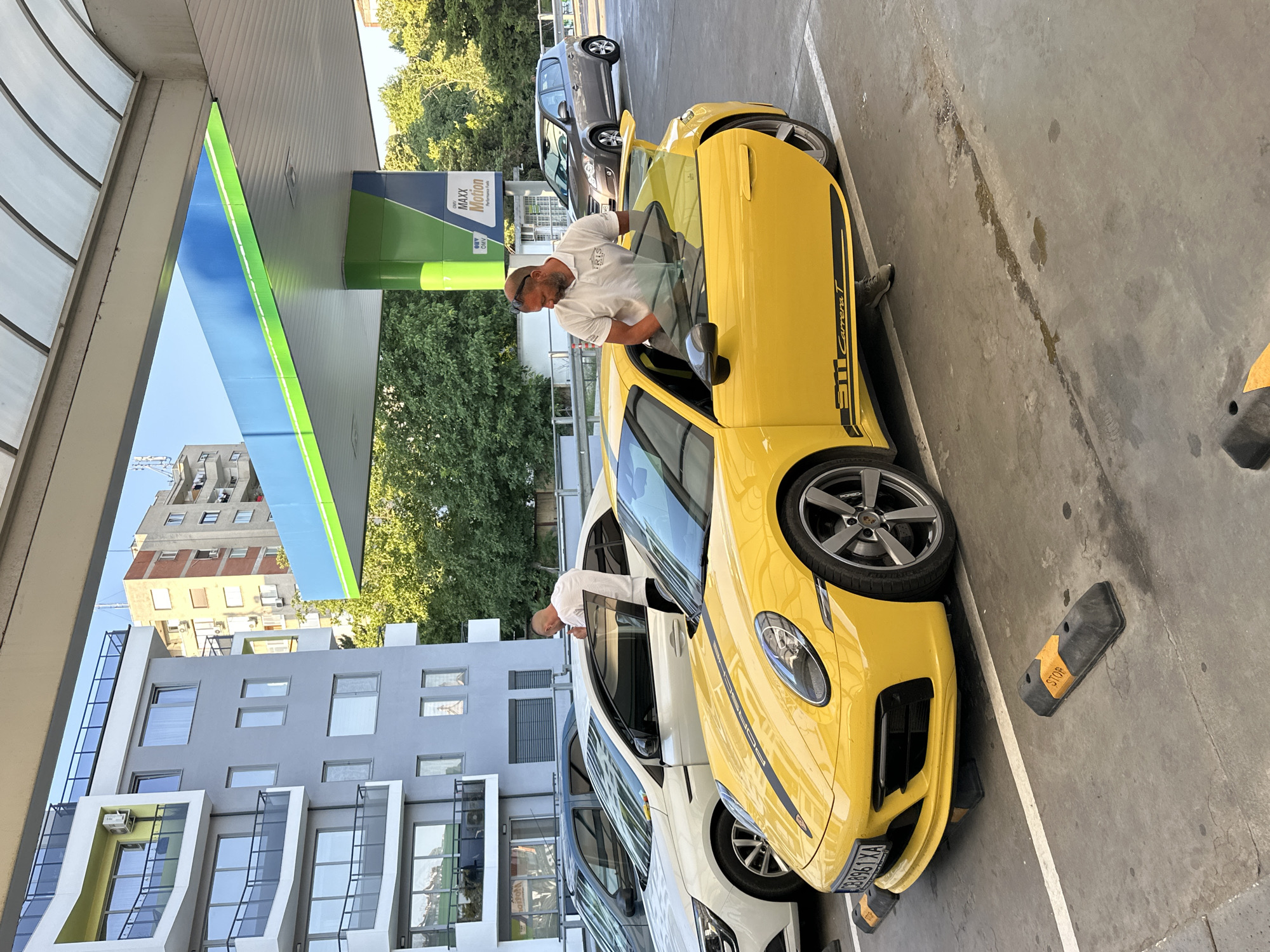 A man is leaning into a bright yellow Porsche 911 Carrera T, which is parked at a gas station. The car has black racing stripes along its side and the "911 Carrera T" text is visible on the side as well. The man is wearing a white polo shirt and sunglasses. Another white car is parked next to the Porsche. In the background, there is a gas station canopy with green and blue signage that reads "OMV MAXX Motion Performance Fuels". Beyond the gas station, there are trees and buildings. The scene appears to be outdoors during the day, with clear skies suggested by the bright lighting. The overall setting is a gas station in Veliko Tarnovo, Bulgaria.