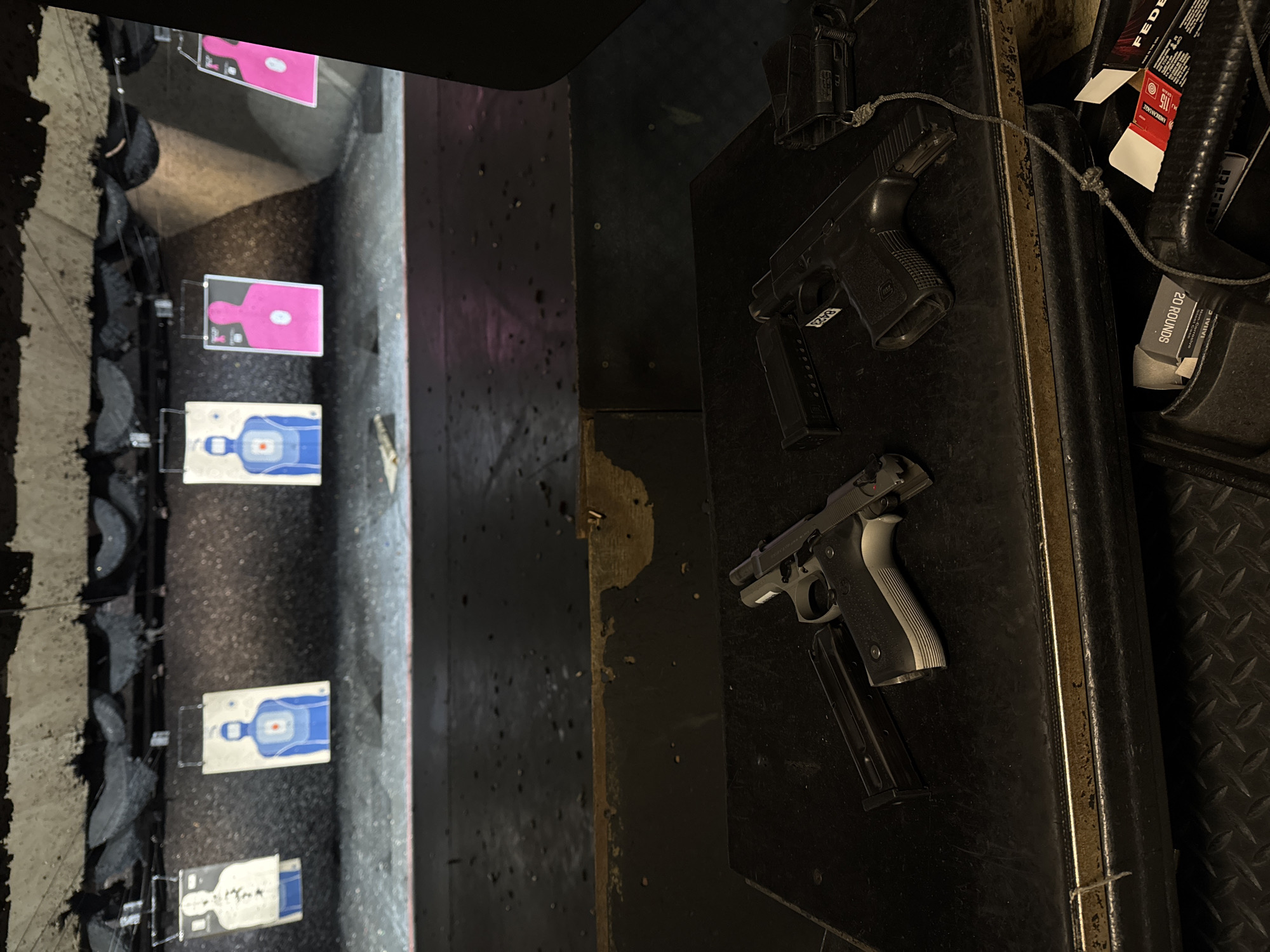 The image shows an indoor shooting range. There are targets hanging in the distance, some with pink human-shaped silhouettes and others with blue human-shaped silhouettes. To the right, two handguns are lying on a black surface, along with ammunition boxes and what appears to be a silencer. The location appears to be in Los Angeles, United States of America. The range shows evidence of prior use with bullet holes in the walls. The presence of firearms, targets, and ammunition indicates an active or recently used shooting range.