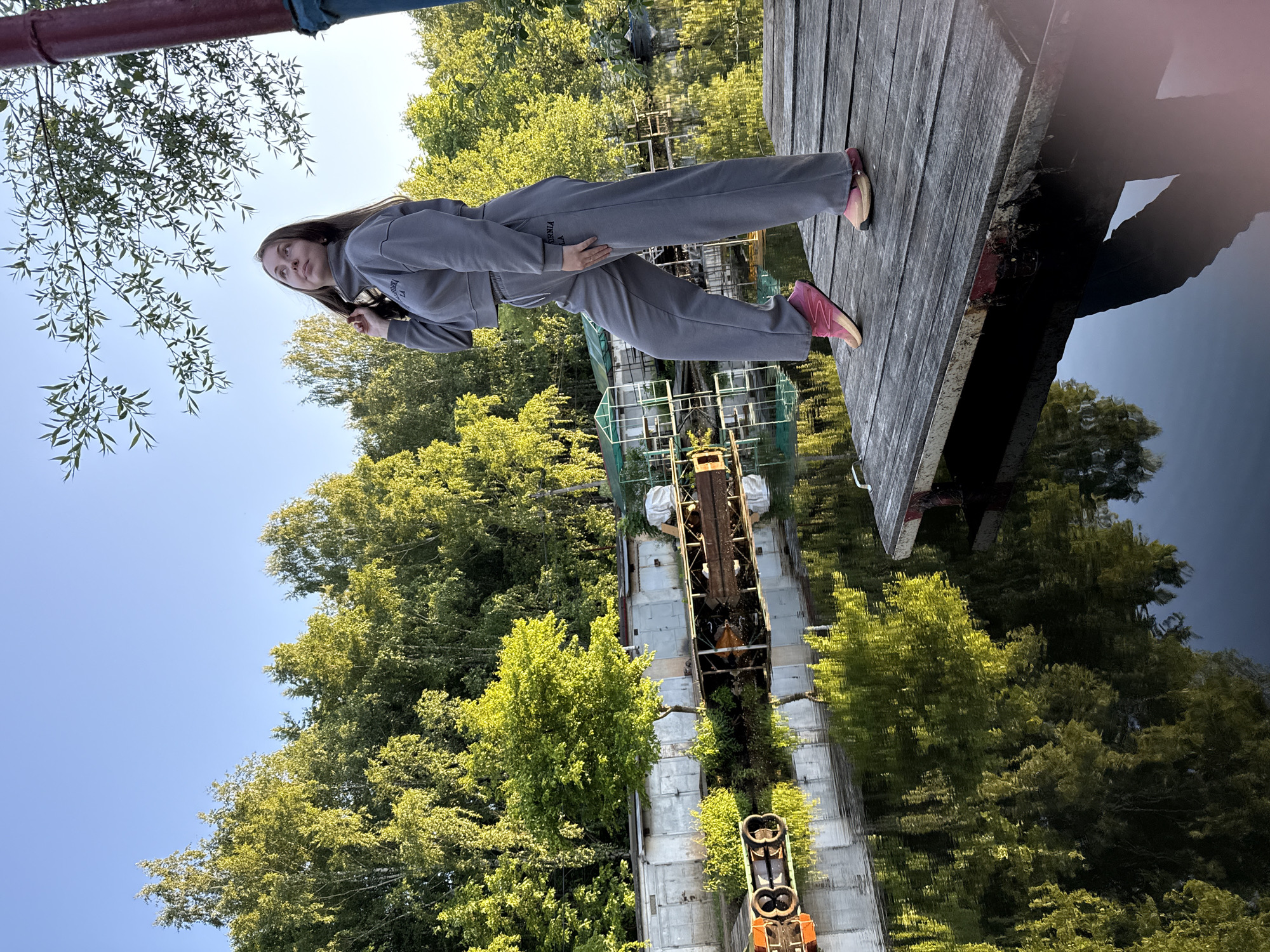 A young woman with long, dark hair is depicted standing on a wooden pier next to a body of water, under a clear blue sky, surrounded by abundant green trees. She is wearing a grey tracksuit, with the words "CALIFORNIA" and "LA" visible in white text on both her sweatshirt and sweatpants. Her sneakers are distinctively pink and beige. She stands with one hand on her hip and the other touching her hair, looking thoughtfully upwards and to her left, away from the camera.
The scene suggests an outdoor setting, possibly a river or canal, flanked by dense foliage. The water is dark and highly reflective, mirroring the trees and sky. In the background, there are industrial or disused structures built into or alongside the water, featuring concrete elements and metal machinery, possibly remnants of an old lock, dam, or port infrastructure. The vibrant green of the trees indicates a lush environment, and the clear blue sky suggests a bright, sunny day. A red pipe or railing is partially visible in the immediate foreground. The overall mood is serene, with a touch of contemplation from the subject.