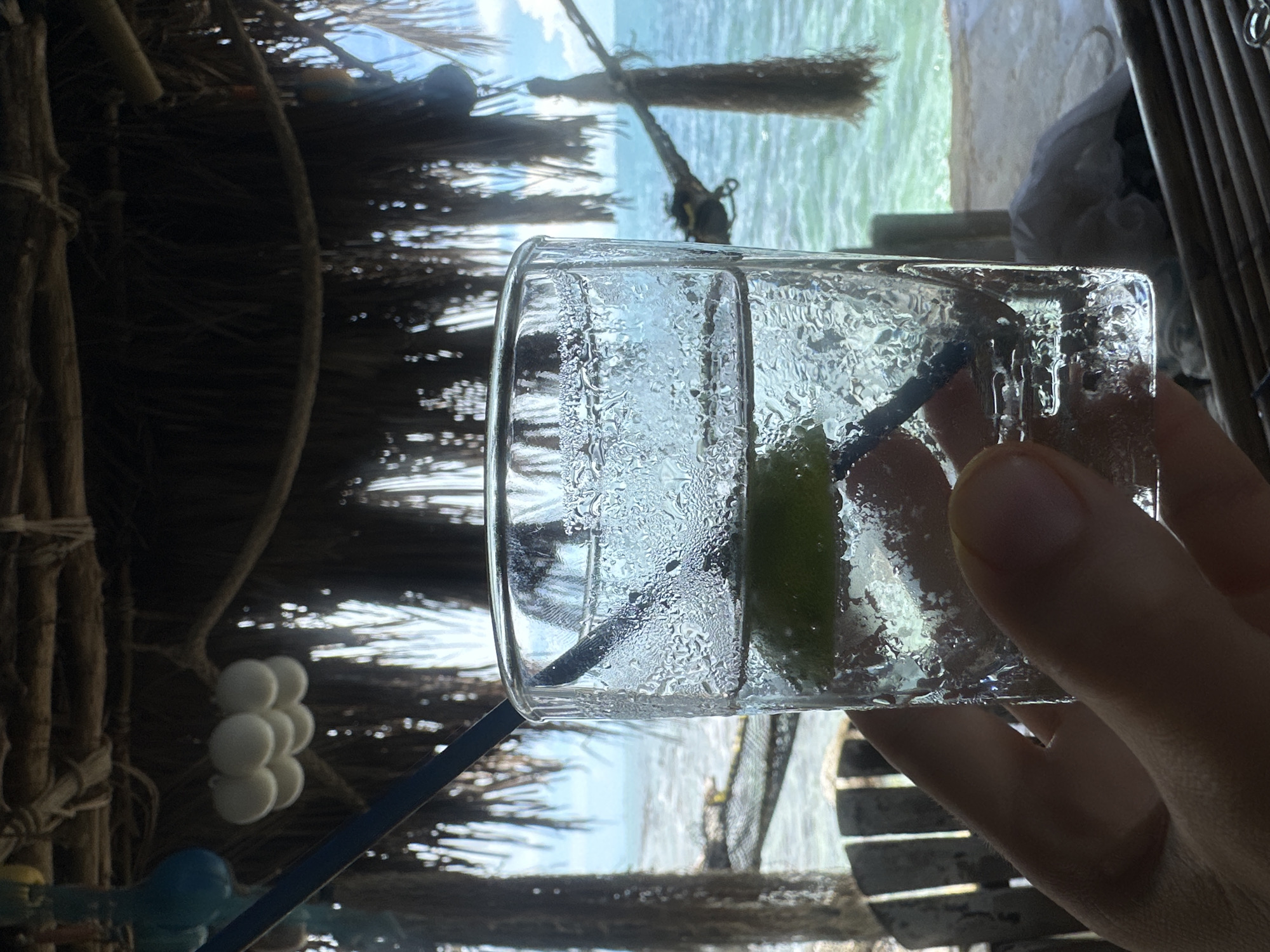 A close-up shot focuses on a condensation-covered glass containing a clear liquid, a slice of lime, and a dark blue straw. The glass is held in a person's hand. In the background, out of focus, is the ocean, a thatched-roof structure, and what appears to be beach chairs. The scene suggests a tropical setting, possibly on Ko Pha Ngan island in Thailand, given the mention of ตำบลเกาะพะงัน. The light is bright, indicating daytime.