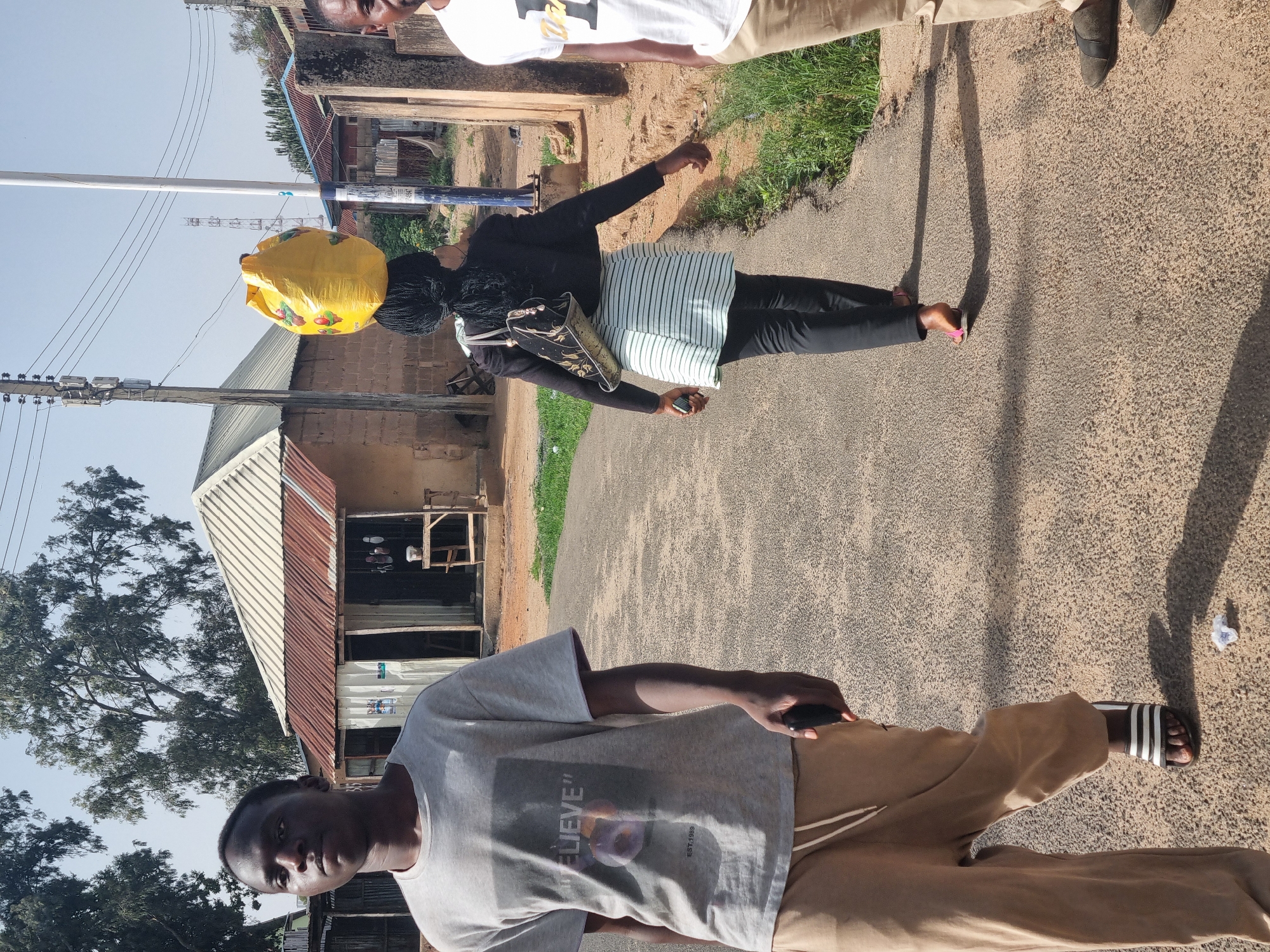 The image shows a street scene in what is likely Jos, Nigeria. There are three people visible: a person in the foreground wearing a grey T-shirt and khaki pants, holding a phone; a woman in the middle ground carrying a large yellow bag on her head and a striped bag on her shoulder, wearing a black top and pants; and part of a third person in the background wearing a white shirt and khaki pants. The woman is walking along a paved road. The environment includes buildings with corrugated metal roofs and utility poles with power lines. The road surface is asphalt, and there is some vegetation along the roadside. A building in the background appears to be a shop or small business.