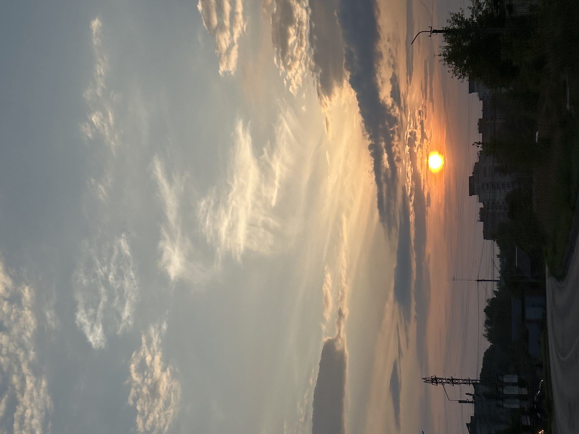 The image captures a sunset over the city of Chaikovsky, Russia. A bright orange sun is visible near the horizon, casting a warm glow across the sky which is filled with scattered clouds ranging from white to dark gray. In the foreground, a curved asphalt road leads the eye into the scene. To the left of the road, there are several low-rise buildings and what appears to be an industrial structure with electrical towers. Further into the scene, taller residential buildings form the city skyline. Silhouetted trees dot the landscape, and a single streetlight stands on the right side of the road. No people are discernible in the image. The overall atmosphere is serene, typical of a late afternoon or early evening in an urban setting.