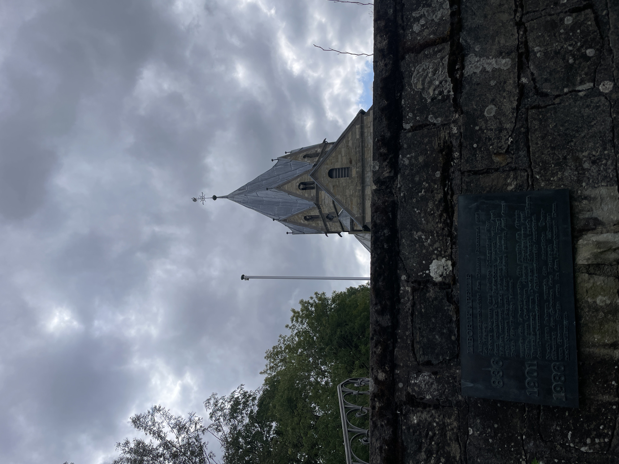 The image shows a stone wall in the foreground with a dark metal plaque attached to it. Above the wall, a church tower with a pointed roof and a weather vane is visible against a cloudy sky. To the left of the tower, trees and a flagpole are present. The plaque on the wall appears to be a historical marker with text and dates. Based on the text on the plaque which includes "Salzkotten", it is identified as the Libori Kapelle in Salzkotten, Germany. The plaque commemorates events related to the chapel, with dates such as 836, 1901, and 1986. The environment appears to be overcast, suggesting a cloudy day. There are no people visible in the image.