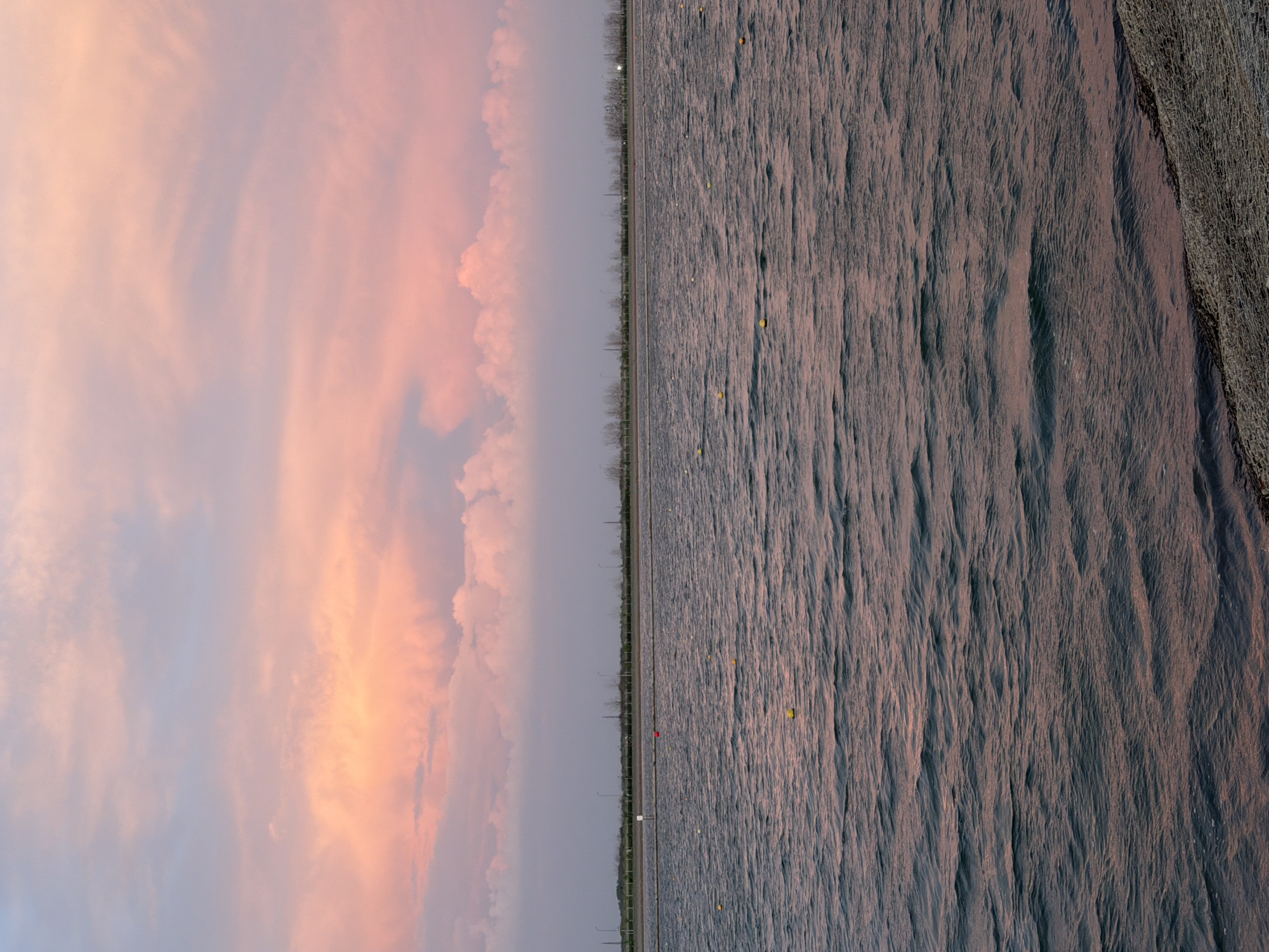 The image depicts a sunset sky with orange and pink clouds on the left side, separated by a road with sparse trees from a body of water with sandbars or shoreline on the right. The location is unknown. The road appears to have occasional light posts. The water shows patterns suggesting ripples or shallow areas. There are no visible people.