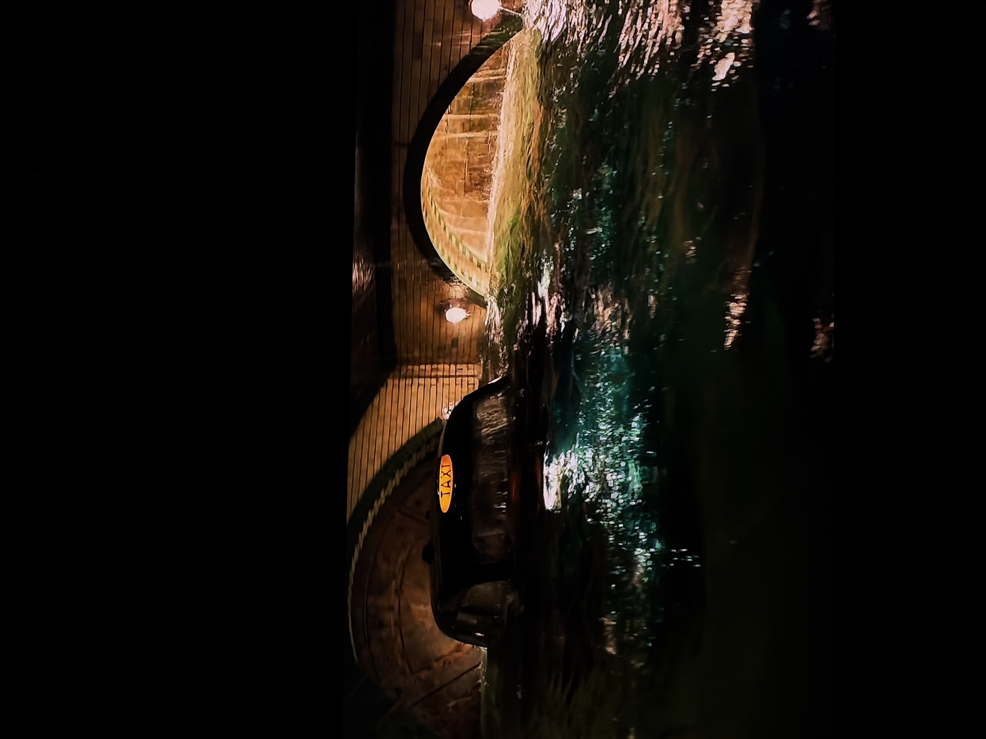 The image captures a nighttime scene featuring a dark taxi cab traversing a watery path beneath an arched structure. The taxi, partially submerged in the water, has a distinctive yellow "TAXI" sign on its roof. The archway, likely an underpass or tunnel, is constructed from what appears to be brick or stone, with vertical wooden paneling on its inner surface. Two warm, glowing light fixtures are mounted within the arch, casting illumination on the structure and the surroundings.
A significant body of water, heavily rippled and reflecting light, occupies the foreground and right side of the frame. The reflections create shimmering patterns of light on the water's surface, indicating artificial light sources either from the taxi or other unseen elements.
The setting is an urban or semi-urban environment at night, suggesting an unusual condition like a flooded street or a canal. No people are visible. The primary visible text is "TAXI" on the car's roof sign. The scene conveys a dark, atmospheric mood, illuminated only by artificial lights, set in Sofiivska Borshchahivka, Ukraine.