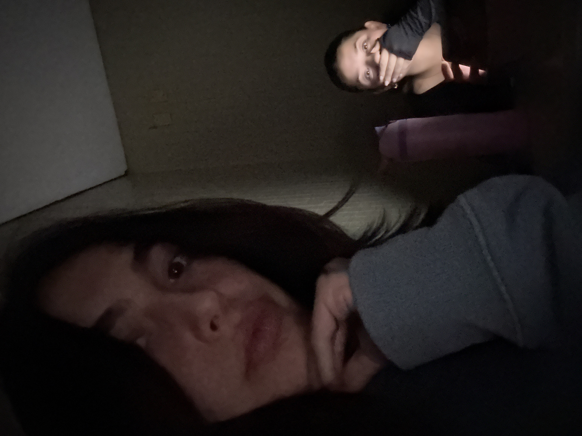The photograph appears to be taken indoors in dim lighting. In the foreground, a person is lying down, facing the camera, with their head resting on a hand covered in a gray hooded sweater. Above and behind this person, another individual is visible, positioned slightly away from the camera. This second person has a hand covering their mouth. A pink cylinder object, possibly a yoga mat or foam roller, is visible in the background behind this person. The environment is dark, and the exact location in the city of Huixquilucan de Degollado, Mexico, is not discernible from the image.