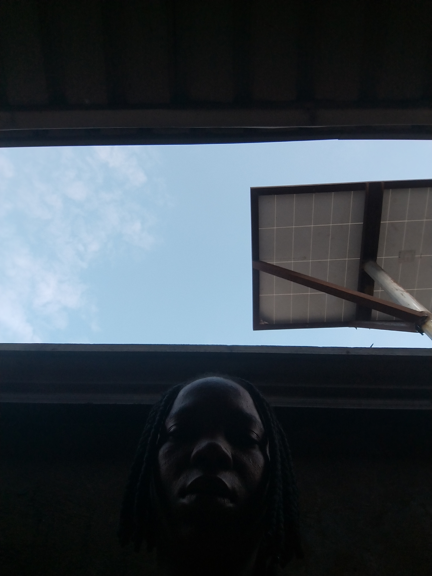 The image is an outdoor, low-angle shot taken from below, looking up at the sky. In the foreground, a person's face is silhouetted against a dark background, with their features obscured by shadow. The person appears to have braided hair. Above the person, a bright blue sky with scattered clouds is visible. To the right of the sky, a solar panel is mounted on a pole, angled towards the sky. The solar panel is framed by dark structures, possibly roofing or awnings, which create a sense of being enclosed or looking through an opening. The overall impression is one of a person looking up from a shaded area towards the sky and a solar panel. The scene is set in Isiehor, Nigeria.