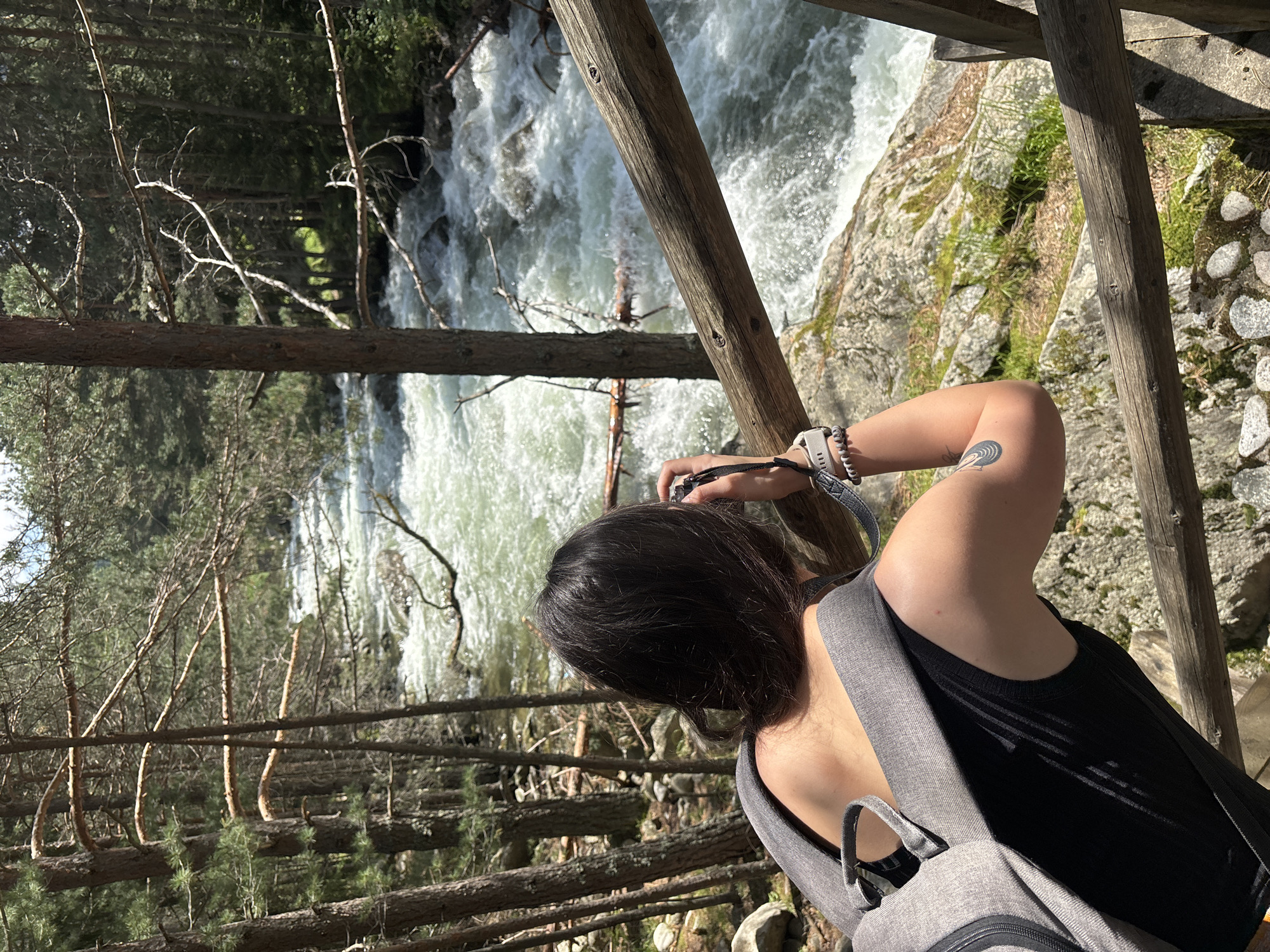 The image captures a person from a slightly elevated perspective, viewed from the upper back and right side, photographing a fast-flowing river in a forested environment, stated to be in Beli Iskar, Bulgaria. The individual has dark hair and is wearing a black sleeveless top and a grey fabric backpack. They hold a compact, dark-colored camera with both hands, aiming it towards the river. A light-colored wristband or smartwatch and a dark beaded bracelet are visible on their right wrist, and a dark, spiral-shaped tattoo is present on their upper right arm.
The river, occupying the mid-ground and background, features significant white rapids and turbulent, light green water, indicating a strong current. Dense coniferous trees with prominent brown trunks and some bare branches line both banks of the river, filling the foreground and background with foliage and dappled sunlight. A rustic wooden railing, composed of two horizontal logs, extends across the lower right portion of the frame, providing a safety barrier. Rocky ground with sparse green vegetation is visible beneath the railing and along the riverbanks.