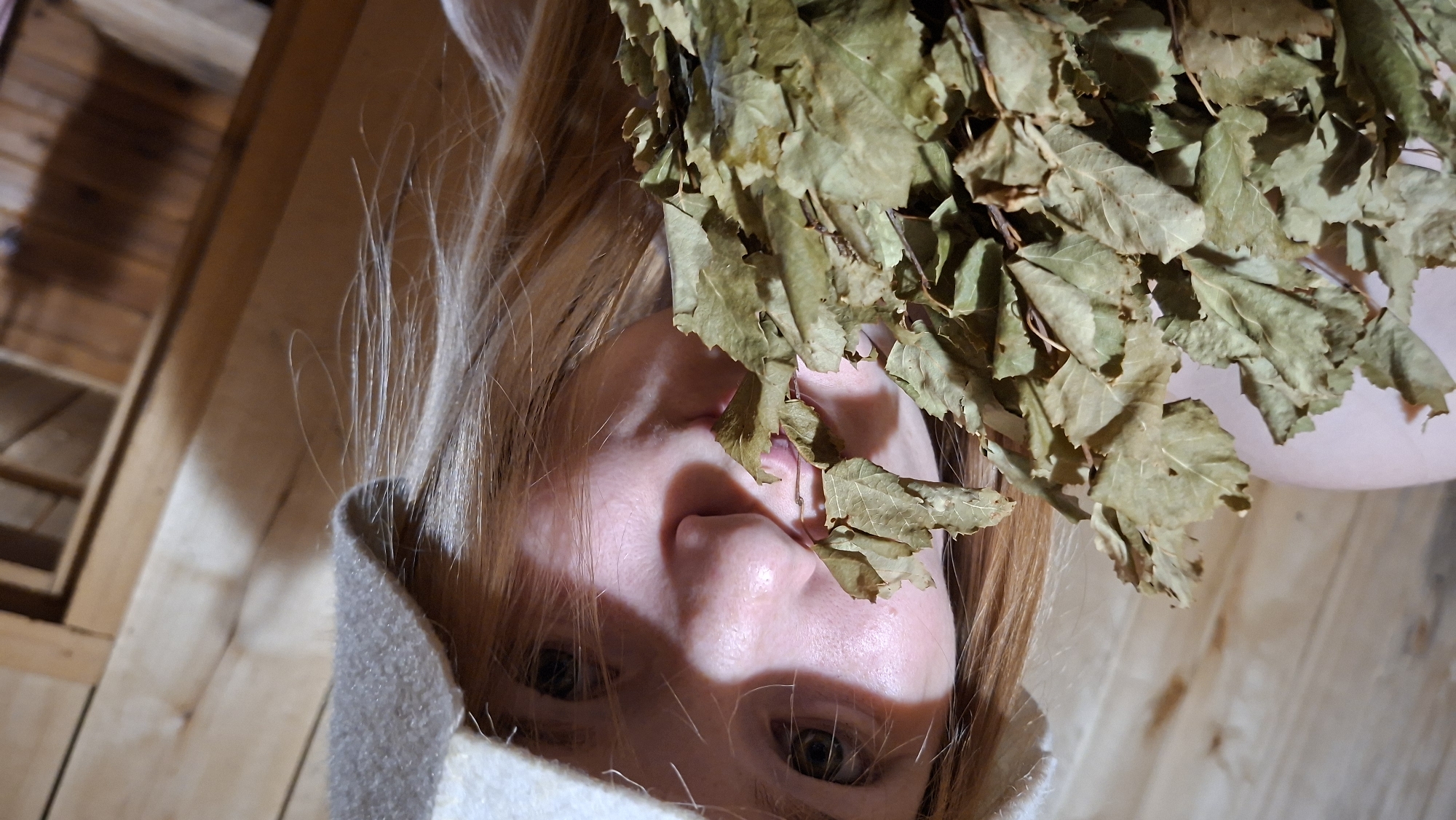 The image shows a close-up of a young woman with blonde hair, wearing a white felt hat, holding a bunch of dried birch branches in front of her face. Her lips are slightly pursed, and she is looking towards the camera. The background consists of wooden walls, suggesting the interior of a sauna or a similar wooden structure. The lighting is warm and somewhat dim, casting shadows on the wooden surfaces. There is no identifiable location within the city of Большой Букор, Russia that can be determined from the visual information provided.