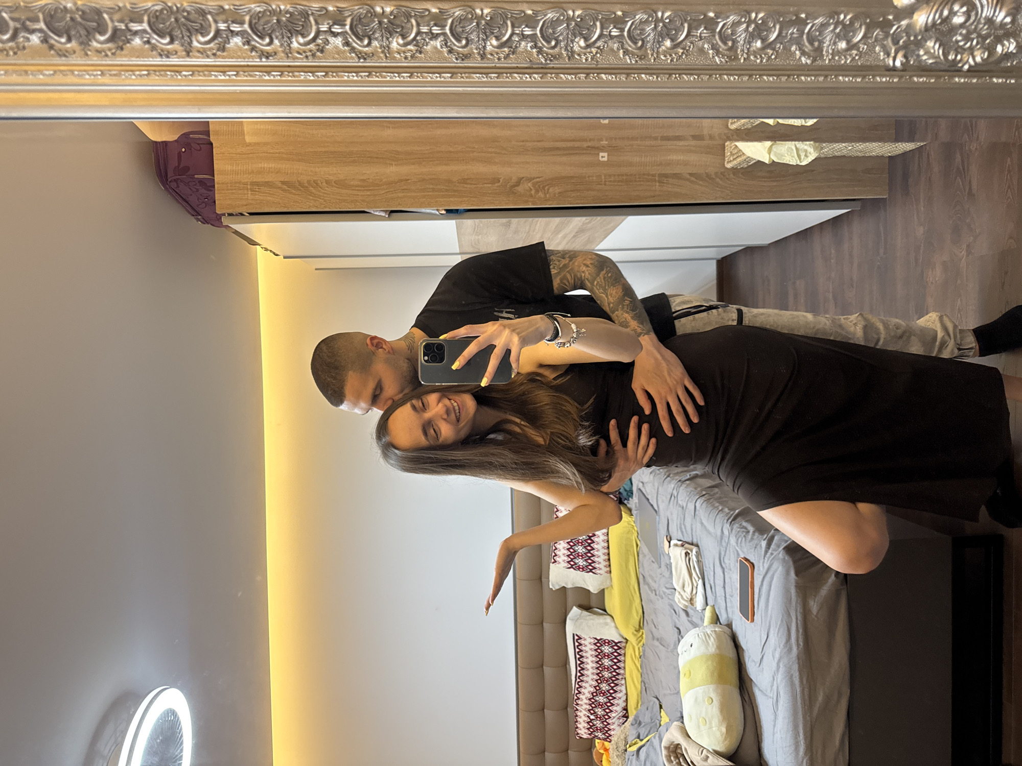 A mirror selfie captures a man and a woman in an indoor setting. The man, with a shaved head and tattooed arm, is wearing a black t-shirt and grey pants. The woman, with long brown hair, is wearing a black dress. They are embracing, with the man holding a smartphone to take the photo. The background reveals a bed covered in a grey sheet with various items on it, including pillows and a phone. A cabinet is visible in the background, along with a wall adorned with a vertical light fixture. A ring light can be seen on the left. Given the style of the interior, the location is likely a residential space in Sofia, Bulgaria.