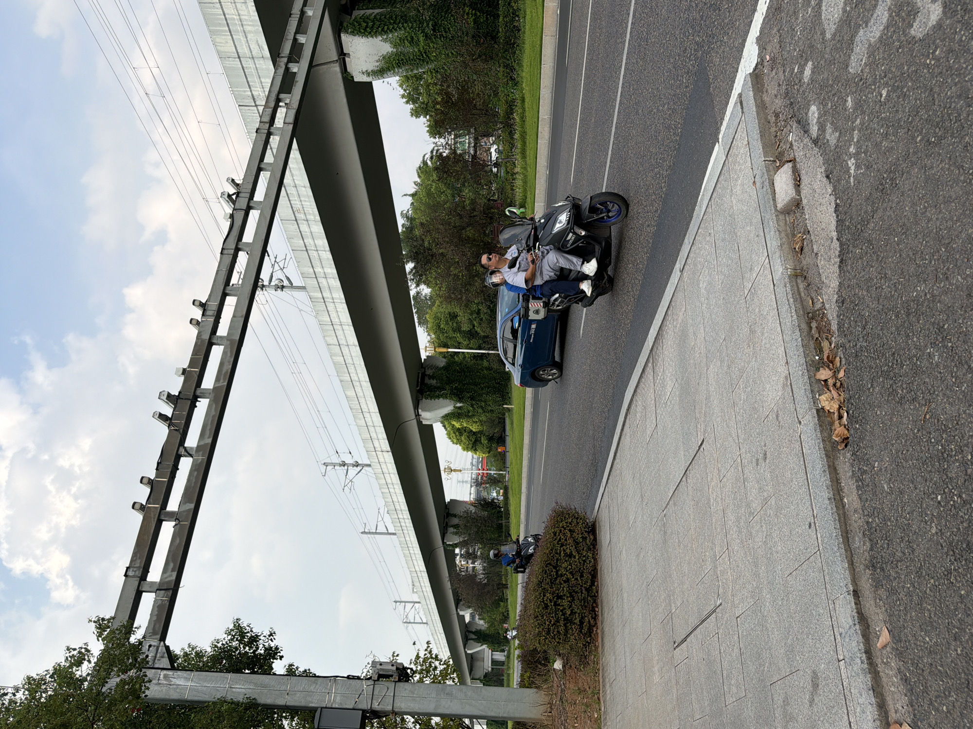 In Jiangjin, China, a street scene unfolds beneath an elevated transportation structure, likely a viaduct for trains or an elevated highway. A light gray paved walkway lines the left side of the image, bordered by a road. On the road, a black scooter with two occupants travels rightward. The driver, a man in a gray shirt and pants, wears sunglasses and a helmet. A passenger, a woman in a blue shirt and black pants, also wears a helmet and sits behind him. A blue sedan is visible further down the road, also moving in the same direction. Overhead, the concrete structure of the viaduct is supported by pillars, some of which are draped in green ivy. Electrical wires are strung beneath the viaduct's structure. The sky is partly cloudy, with patches of blue visible through white clouds. Trees and bushes are present on both sides of the road, with the bushes on the left side appearing meticulously trimmed. A metal pole with electrical equipment is positioned on the far left of the image, near a large tree. Fallen leaves are scattered on the walkway and the edge of the road.