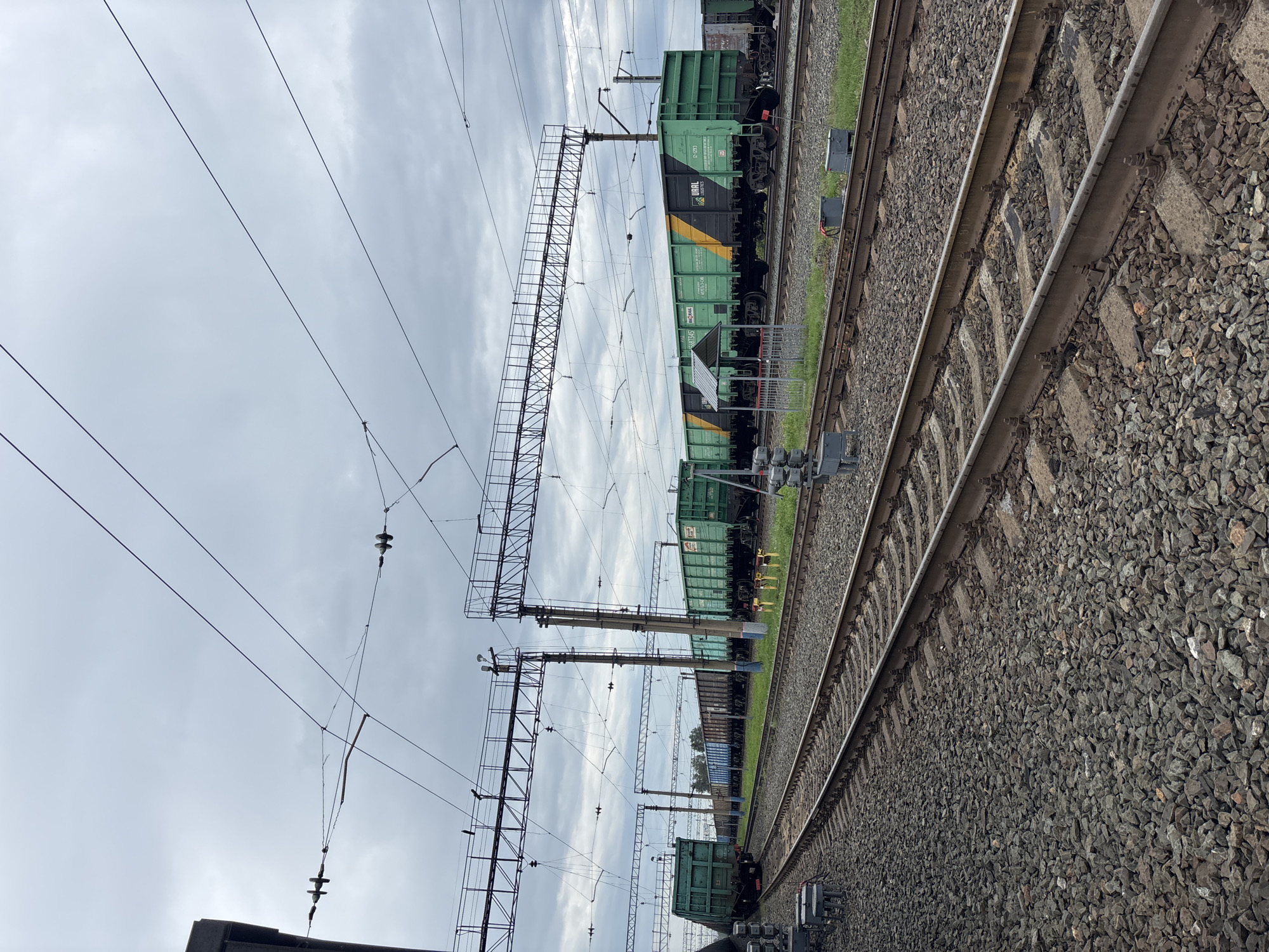 A train yard is pictured under a cloudy sky in Borodino, Russia. The image features several train tracks running parallel to each other, covered in dark grey gravel. Multiple freight cars, primarily green with some yellow and black accents, are parked on the tracks. Overhead, a complex network of wires and support structures for an overhead catenary system is visible. The environment appears to be an industrial area, with no people or animals present in the frame. The overall scene depicts a static, functional setting of a working train yard.