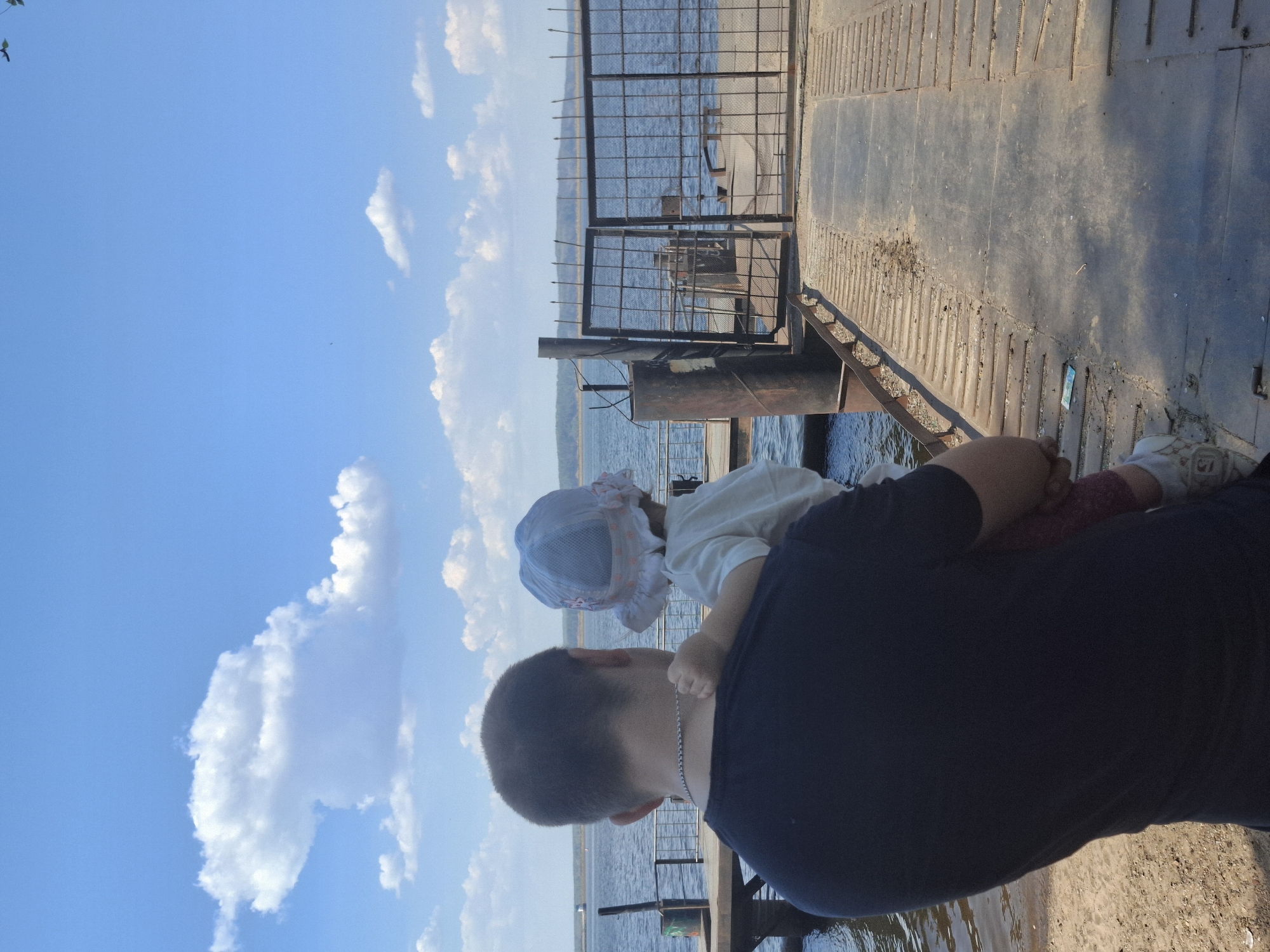 A man holds a young child in his arms while standing on a pier in Chaikovsky, Russia. The man has short hair and a silver chain around his neck. The child is wearing a sun hat. The pier has a metal gate at the end, overlooking a body of water, presumably the Kama River, as Chaikovsky is located on its banks. The sky is blue with scattered clouds.