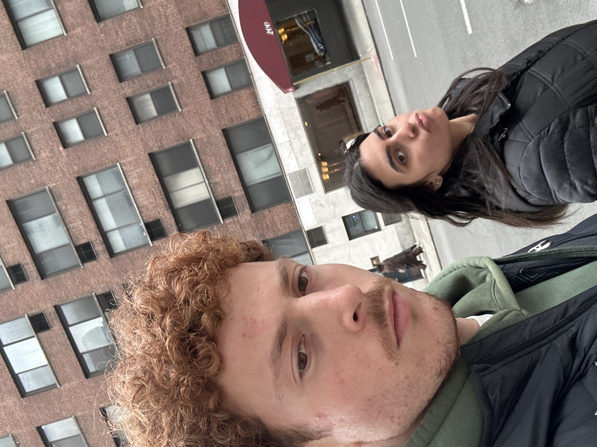A slightly tilted photograph captures two individuals in the foreground and midground of an urban street scene. The primary location is 200 East 61st Street, New York, NY, USA, indicated by the number "200" on a red arched awning visible on the right side of the background.
The foreground features a light-skinned male with reddish-brown curly hair, light mustache, and stubble, looking directly at the camera with a neutral expression. He wears a dark green hooded sweatshirt beneath a black quilted jacket. His face and upper chest occupy the left and central portion of the frame.
To his right and slightly behind him is a light-skinned female with long dark hair, also looking towards the camera, with her lips noticeably pursed in a "duck face" expression. She is wearing a black quilted jacket.
The background consists of a multi-story building featuring a red brick facade with numerous rectangular windows on its upper floors. The lower section of the building has a lighter-colored marble or stone facade with larger windows. A faint reflection of a blue vehicle is visible in a ground-level window beneath the red awning. A blurred figure in a dark coat walks on the sidewalk in the distant background to the right. The asphalt street with white markings is visible in the lower right corner. The scene is illuminated by diffused daylight, and the street appears somewhat damp.