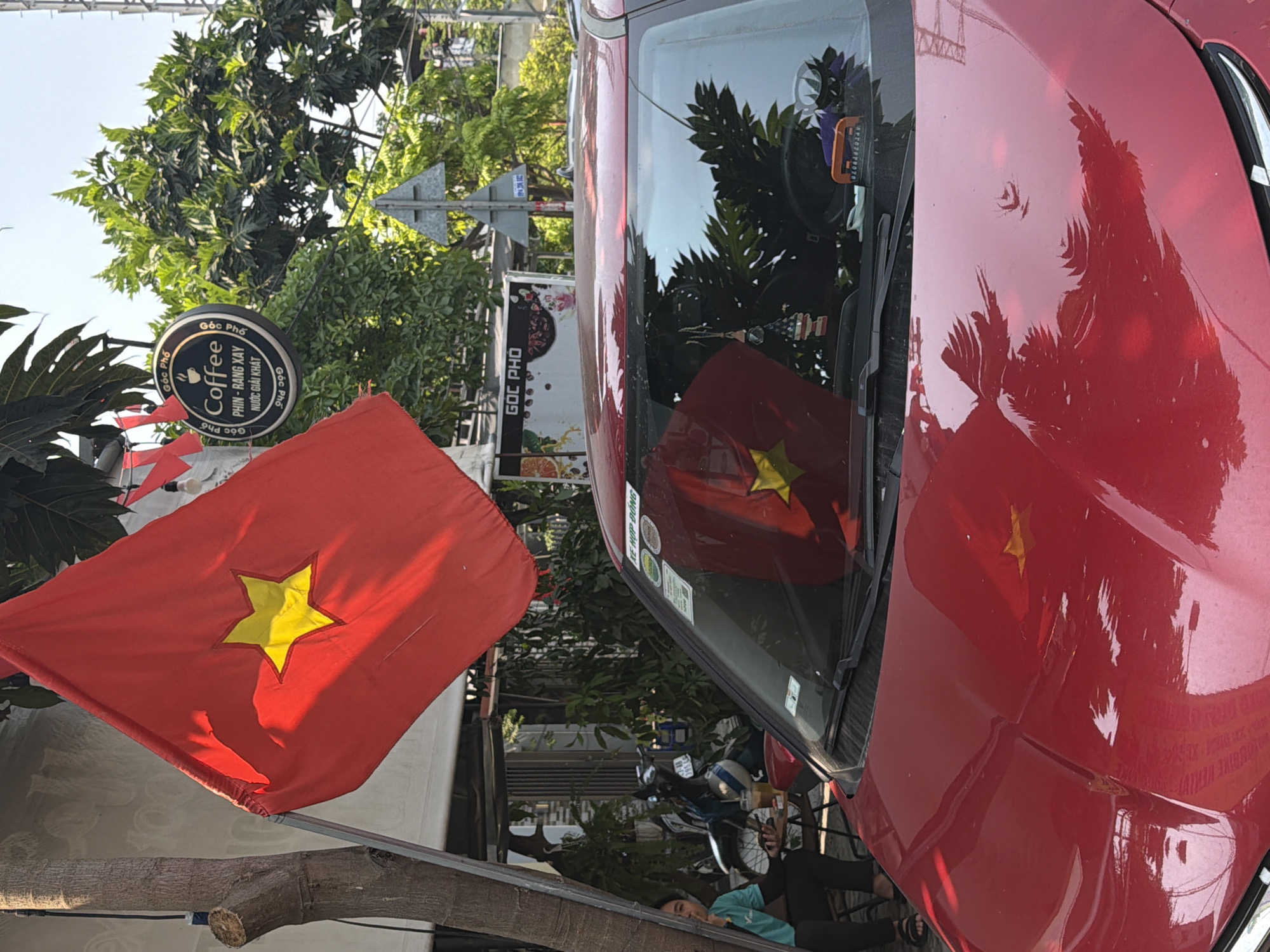 A tilted, medium-shot photo captures a street scene in Đà Nẵng, Vietnam. The focal point is a red car with a Vietnamese flag’s reflection on its hood and windscreen. A real Vietnamese flag flies prominently on the left side of the frame. Behind the car, there is a street with motorbikes, and a person lying down. The "Góc Phố" coffee shop is visible, indicated by signs in the background. The image includes trees and architectural elements.