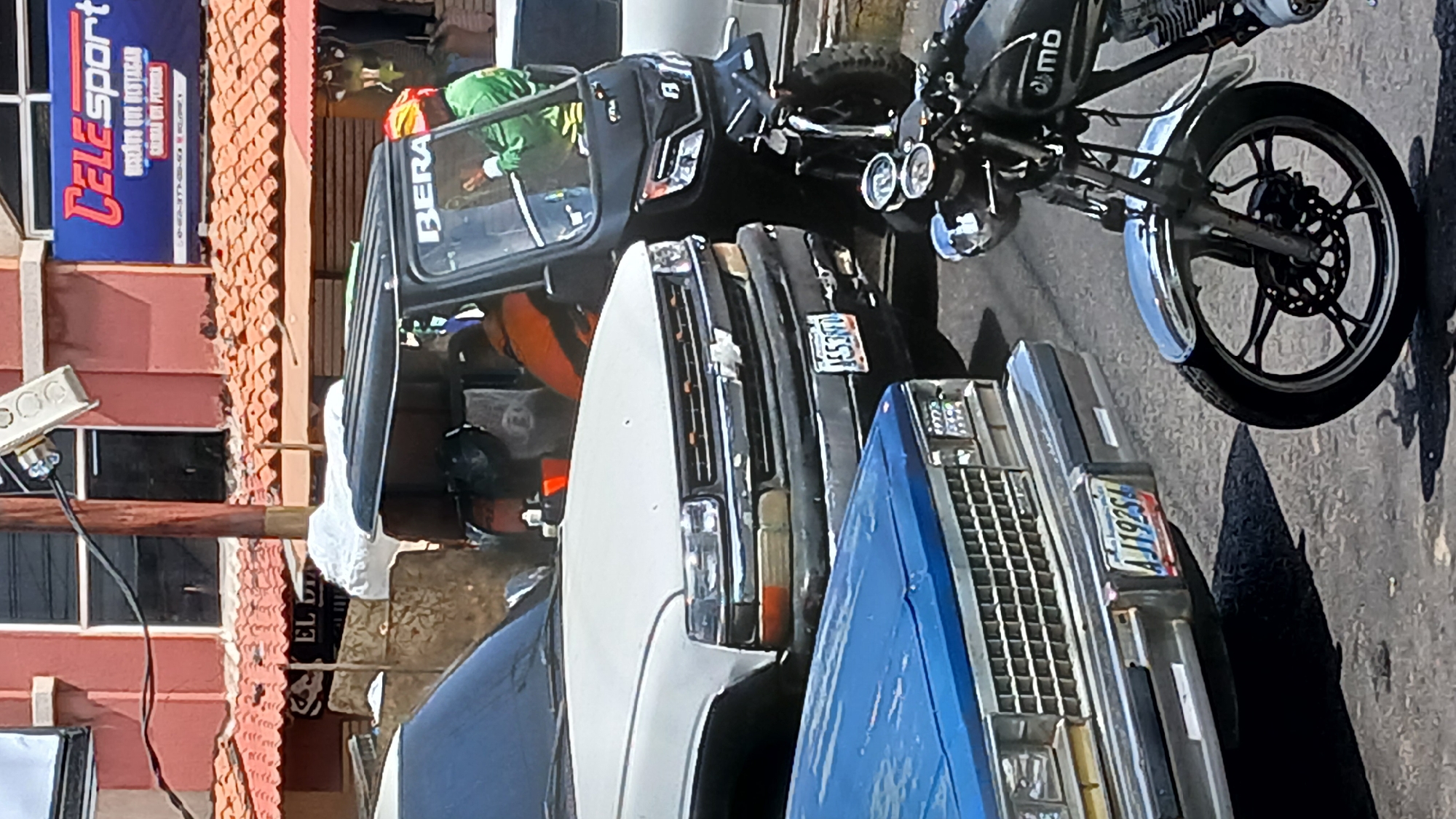 The image shows a street scene in Puerto La Cruz, Venezuela. A "BERA" brand motor tricycle taxi is positioned adjacent to several vehicles. The vehicles include a white pickup truck with the partial license plate "1ES39D" and a blue vehicle with a partially visible license plate "AJJ1925." Parked adjacent to the tricycle is a motorcycle brand "MD." The surroundings show buildings and signage, one reading "TeleSport." The environment appears to be an urban street with parked vehicles. There is a person visible inside the tricycle, wearing green and orange clothing.