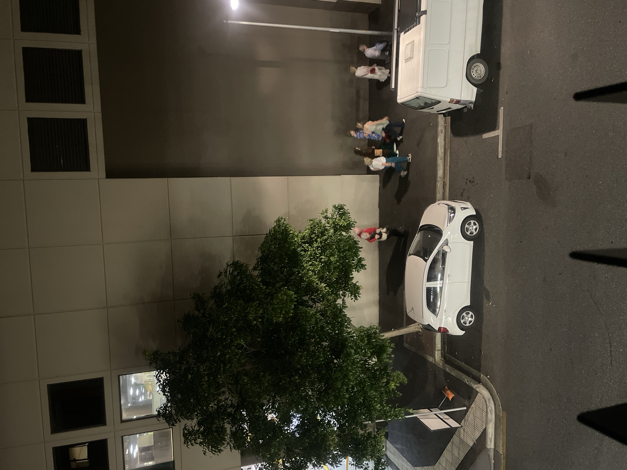 The image shows a nighttime street scene with a building in the background, a tree, and two vehicles. A white hatchback car is parked on the right side of the street. A white van is parked to the right of the hatchback. Several people are walking on the sidewalk in front of the building. The building appears to be a modern structure with windows and vents. The street is paved and appears to be in good condition. The lighting is provided by a streetlamp and interior lighting from the building.
