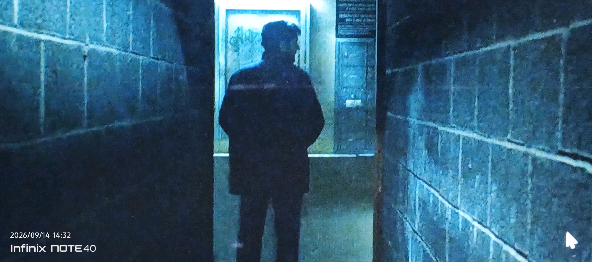The image shows a silhouetted figure standing in a narrow alleyway at night. The figure is facing away from the camera and appears to be wearing a dark coat and pants. The alleyway is lit by a bluish light, possibly from a neon sign or streetlamp. The walls of the alleyway are made of brick and appear to be wet, suggesting that it may have rained recently. There is a sign on the wall to the right of the figure, but the text is unreadable. The bottom left corner of the image displays the date "2026/09/14 14:32" and "Infinix NOTE 40". There is no information in the image or its context to suggest a location in Muyan, Uzbekistan.